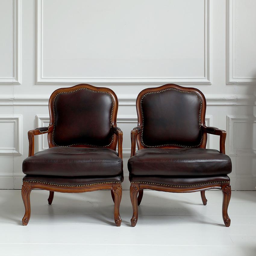 A pair of Louis XV-style fauteuils exuding sophistication and antique charm. Upholstered in dark brown leather with nail head trim outlining the leather edges, which emphasise the elegant contours of the frames. Cabriole legs and shaped aprons