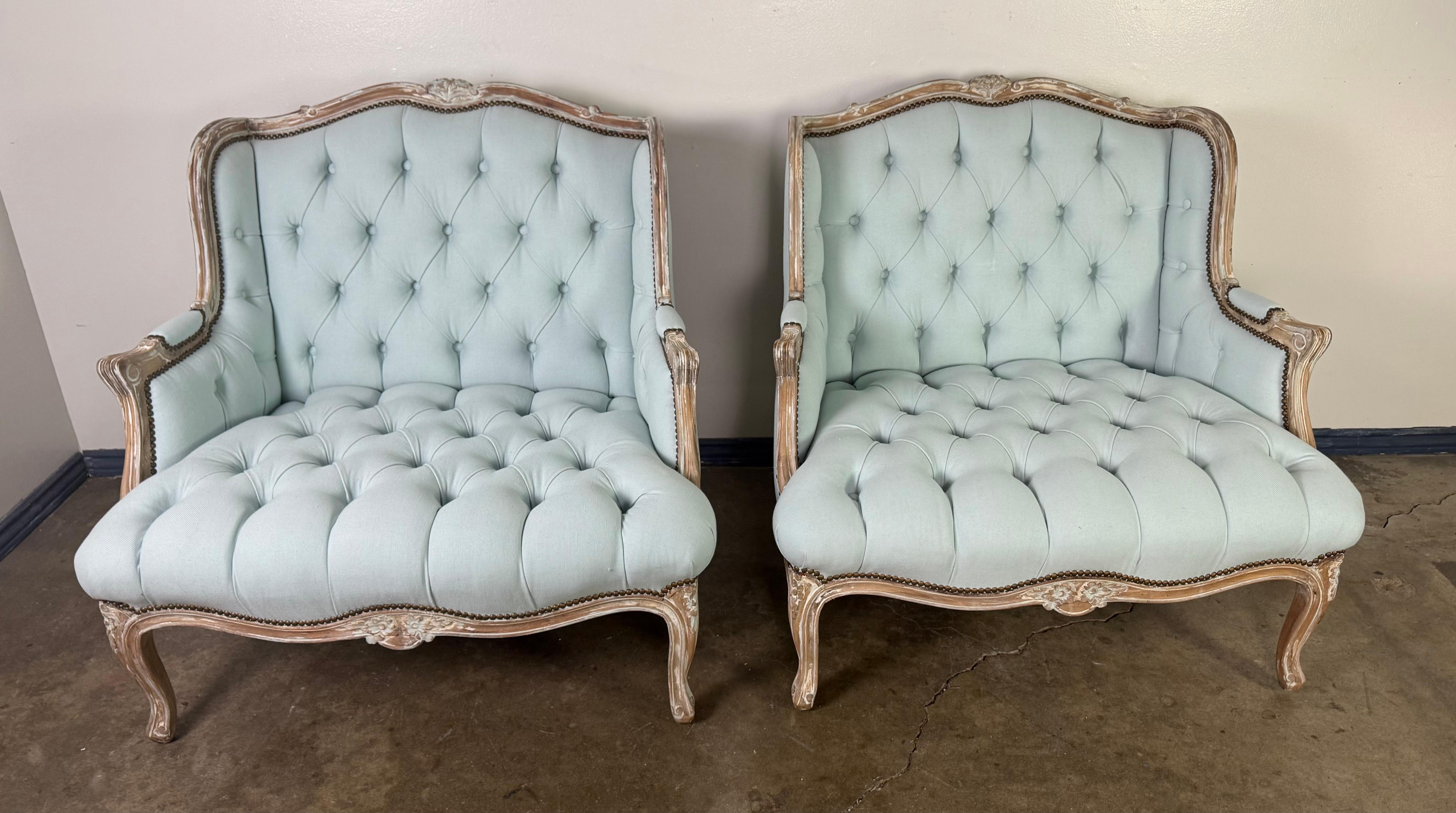 Elegante pareja de sillas Bergère de estilo Luis XV con armazón de madera maciza tallada y un encantador acabado envejecido. Recién tapizadas en un suave lino de color azul pálido natural, las sillas están bellamente mechonadas a lo largo del