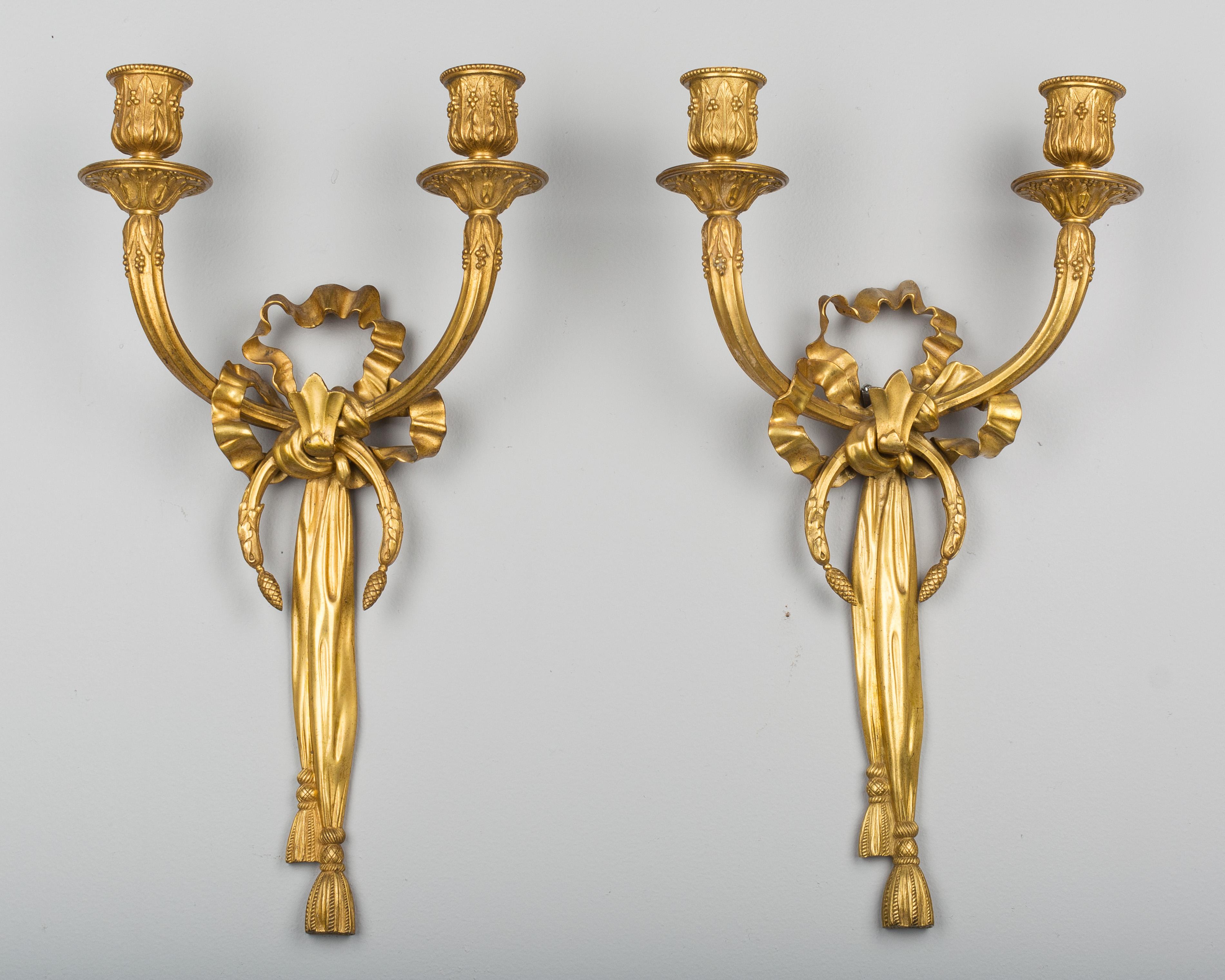 A pair of Louis XV style two-light gilt bronze candle sconces. Ribbon form with tassels and delicate foliate and floral details. Not wired. The bracket on the back of one of the sconces has a welded repair.
   