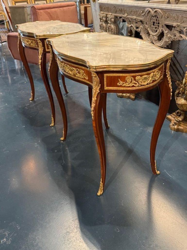 The product is a set of two Louis XV style mahogany and gilt bronze tables. These tables have intricately designed bronze handles and are finished in a luxurious mahogany wood with a beautiful inset marble top.  The elegant and ornate design of