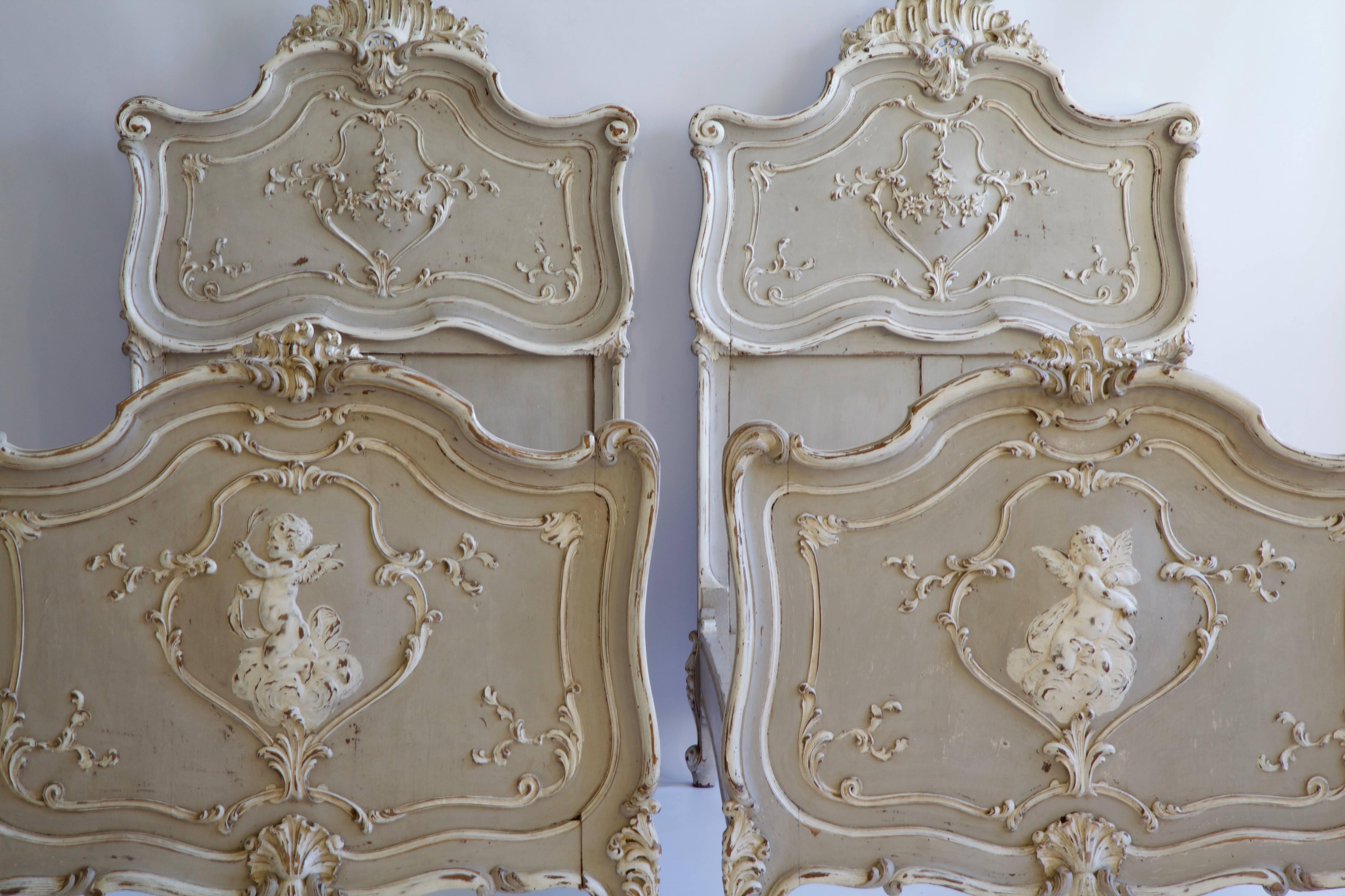 Pair of Louis XV style beds, circa late 19th century. Hand-carved with deeply carved cartouche designs and cherubs as the central theme on the footboards. Finished in the original polychrome paintwork of French grey with Ecru highlights.