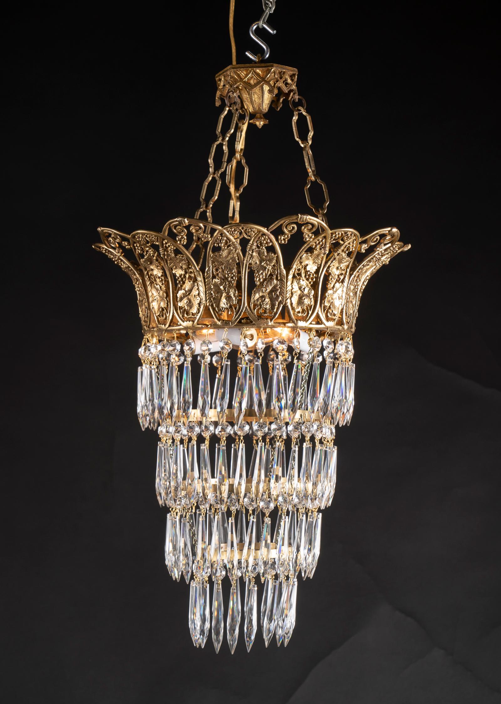 This beautiful pair of Louis XVI style chandeliers features four spherical tiers of crystals suspended from a detailed bronze crown at top. The crown carries a grape motif in reference to Bacchus the god of wine, and dates back to the late 19th