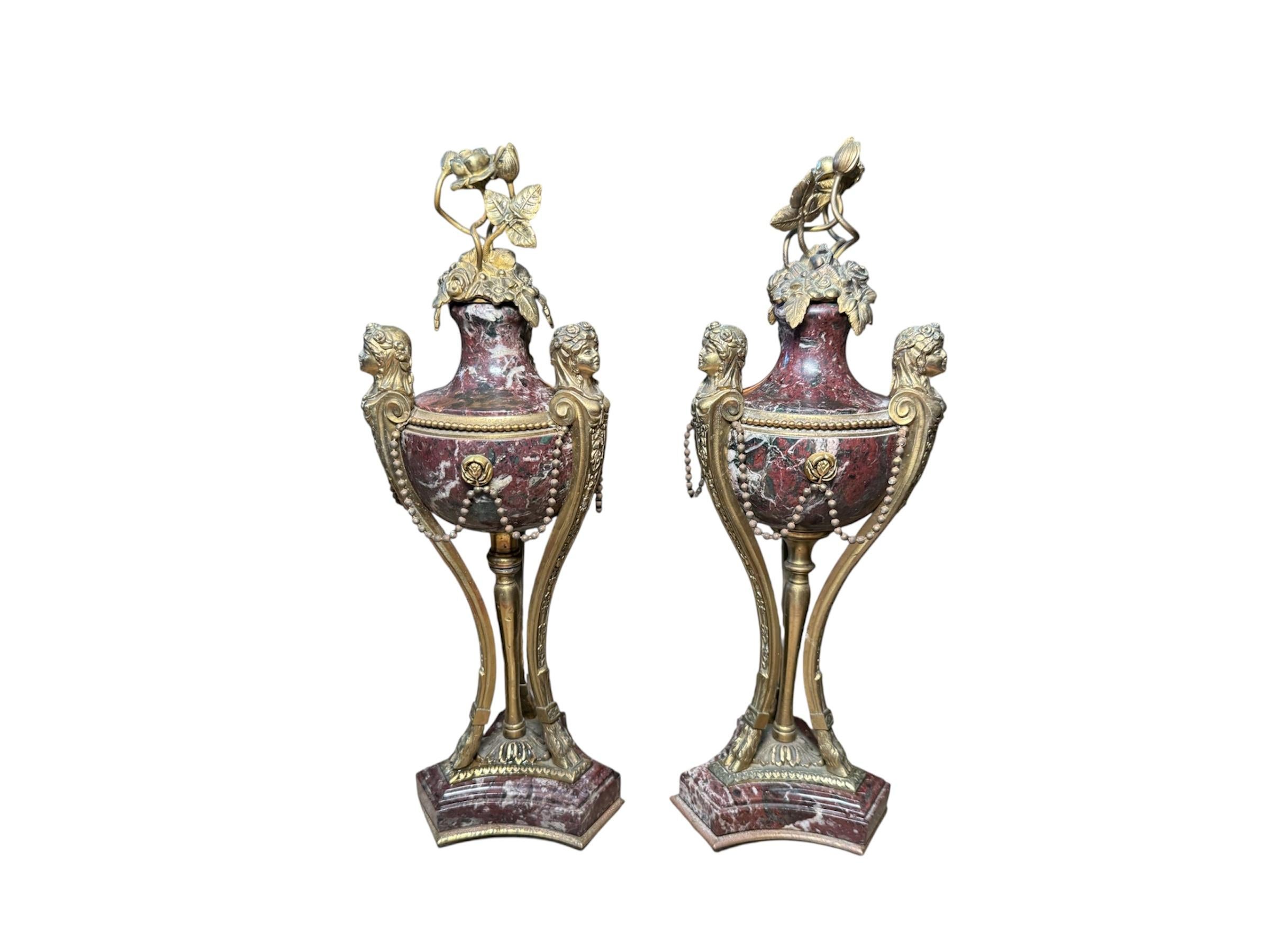 Questo è un paio di Cassolette. Raffigura una ciotola rotonda in marmo di colore bordeaux senza coperchio sostenuta da tre gambe di busti di fanciulle in bronzo incise con fiori e terminanti con zoccoli di ungulati che sono sostenuti da una base