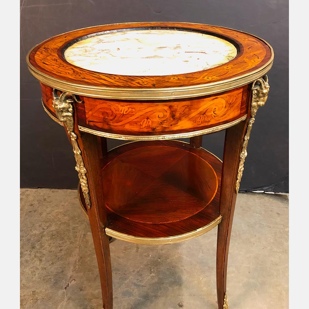 A pair of Italian Louis XVI style marquetry inlaid round pedestal side tables with Carrara marble inset tops, bronze trim and bronze ram's head mounts and hoof foot sabot, with a shelf stretcher base.