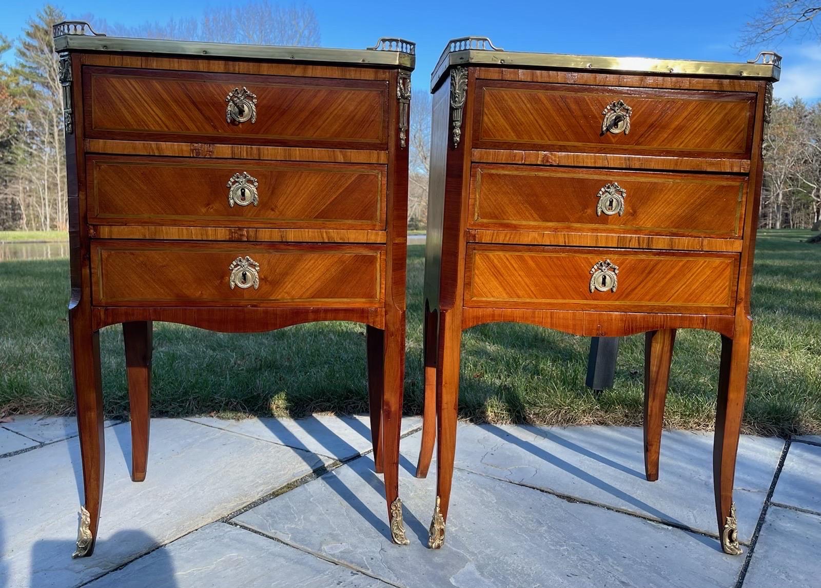 An elegant Pair of Louis XVI style Tulip-Wood, Olive- and Kingwood Veneered Side Tables. The light earthtone Marble Top‘s are in excellent condition and are framed with a solid brass Gallery, open in the front. Three drawers are identical in size