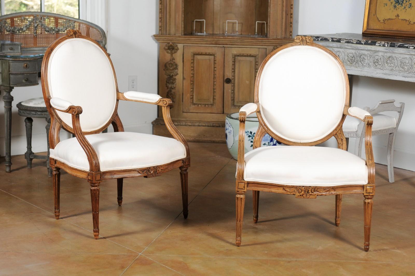A pair of French Louis XVI style oval back fauteuils from the 19th century, with carved crest, scrolling arms and upholstery. Born in France during the 19th century, each of this pair of armchairs features an oval back topped with a floral carved