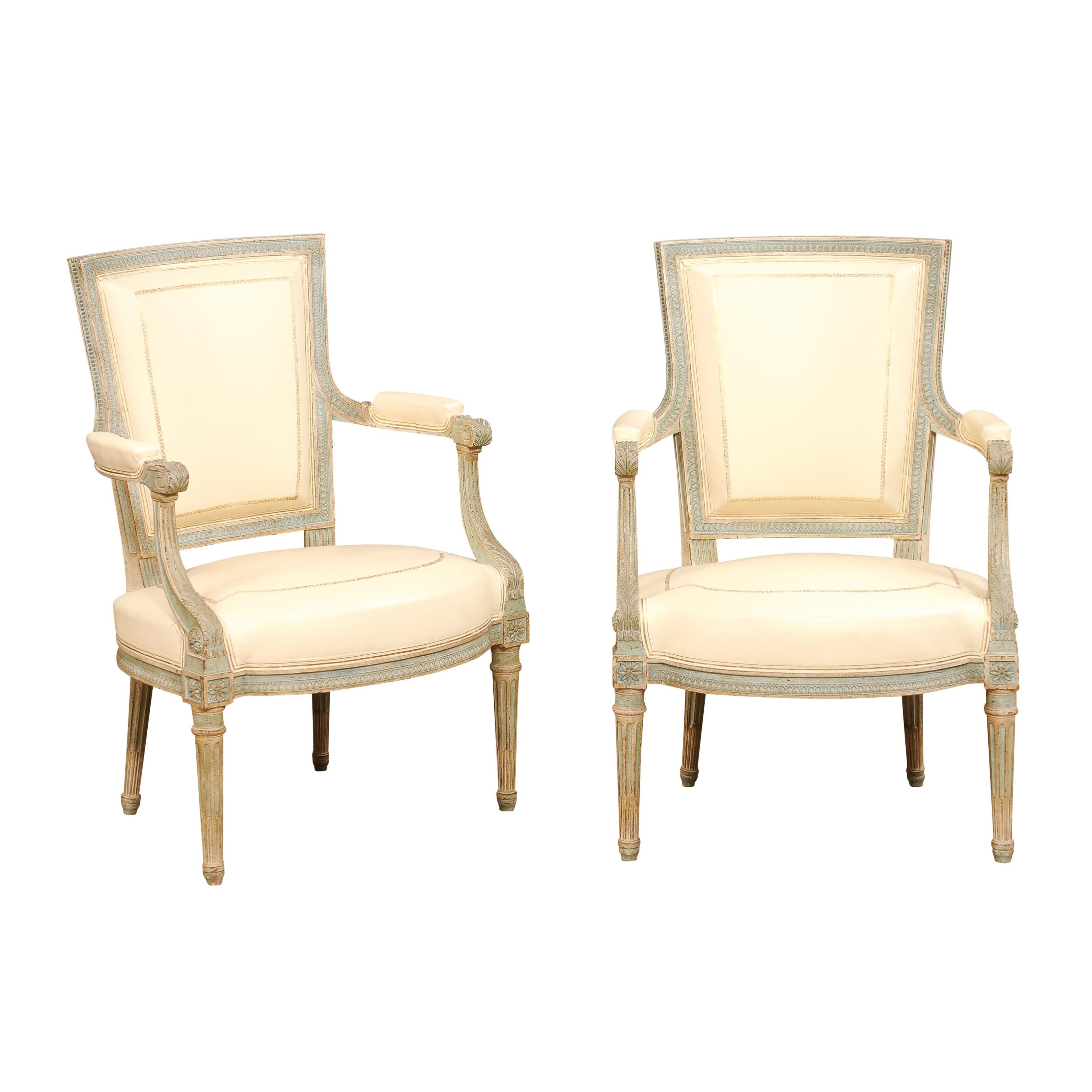 A pair of French Louis XVI style painted armchairs from the 20th century, with white leather upholstery, carved motifs and fluted legs. Emanating elegance and timeless design, this pair of French Louis XVI style painted armchairs from the 20th