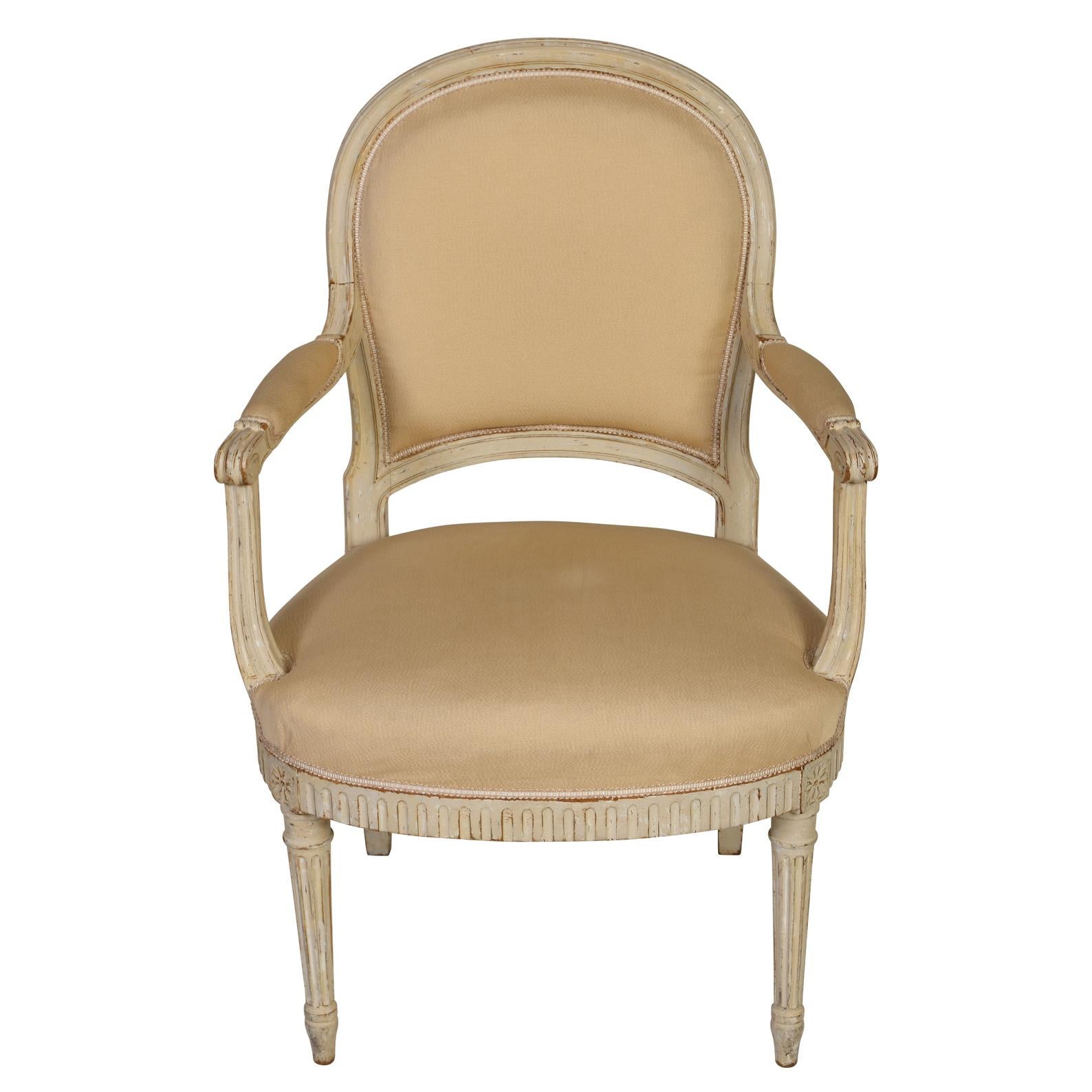 A pair of Louis XVI style fauteuils painted antique white and upholstered in a neutral cream fabric. Curved back and scroll arms with padded arm rests and fluted, carved seat apron with flower detail and fluted carved legs.