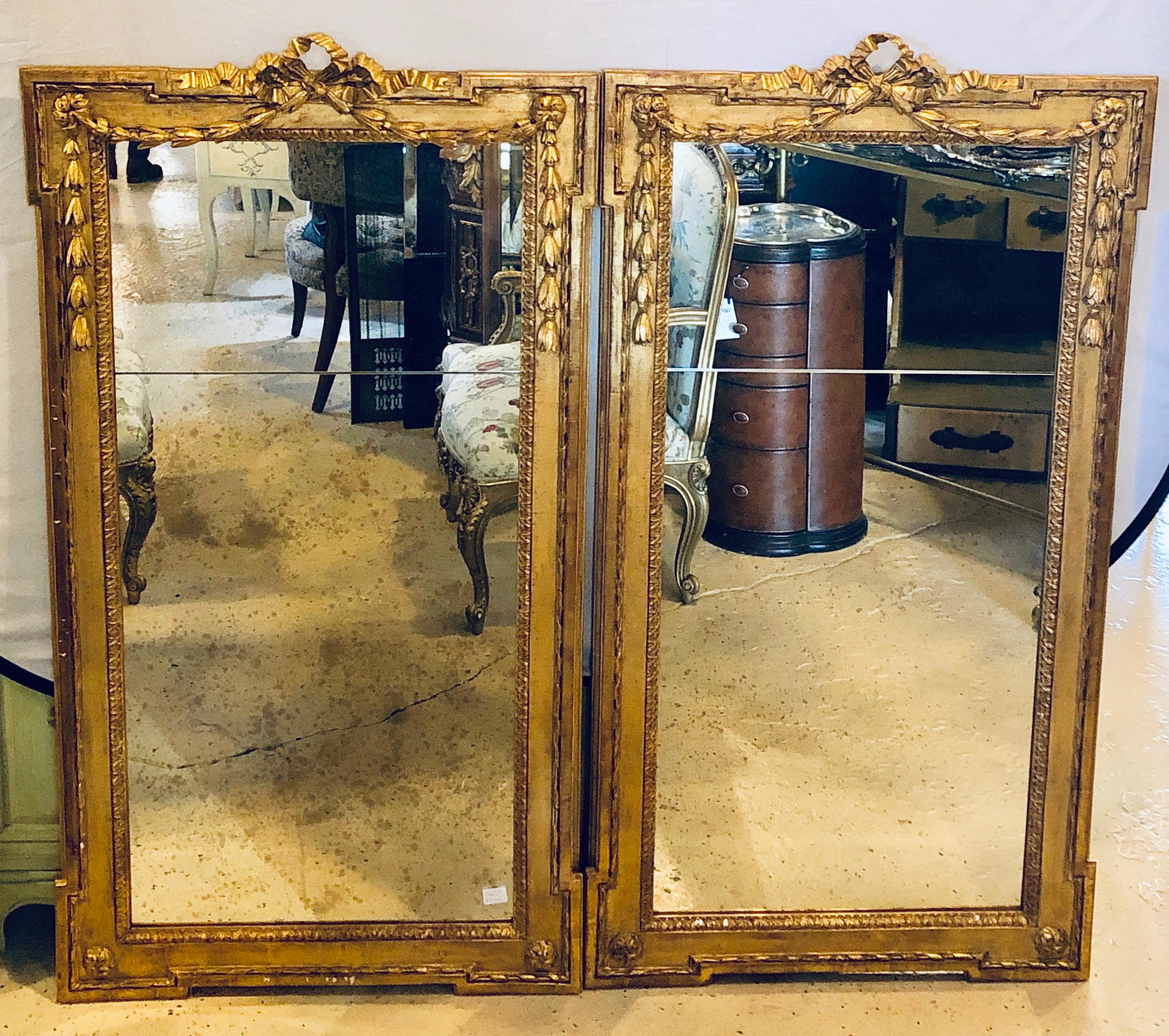 Pair of large and impressive Louis XVI style carved gilt gold wooden wall or console/floor mirrors. Each rectangular frame in a carved gilt gold finish terminating in carved wreath and ribbon with bow design. The clear center frame mirror flanked by