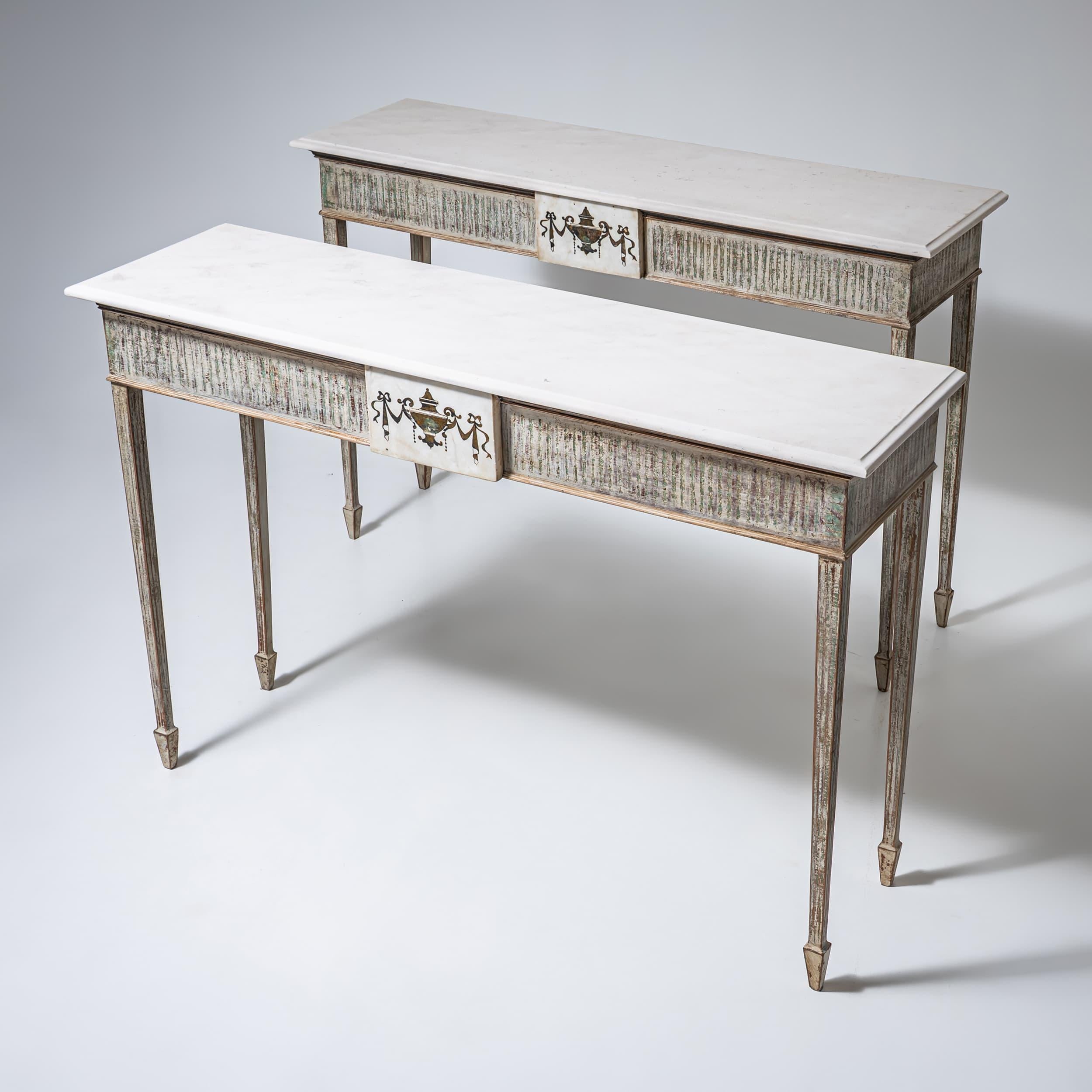 Pair of consoles on tall tapered legs with fluted aprons and white cast-stone tops. The fronts are accented with urn motifs executed in pietra dura technique. The light beige paint finish is newly applied and given an antique patina.