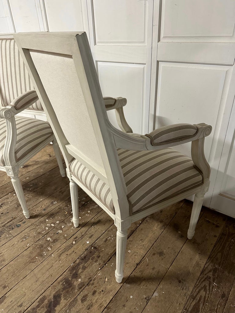Pair of stylish vintage French Louis XVI style arm chairs, upholstered in beige & tan striped linen on the backs, arms and seats with a solid fabric on the backs. These chairs have frames painted in soft shade of French white. Chairs are perfect to
