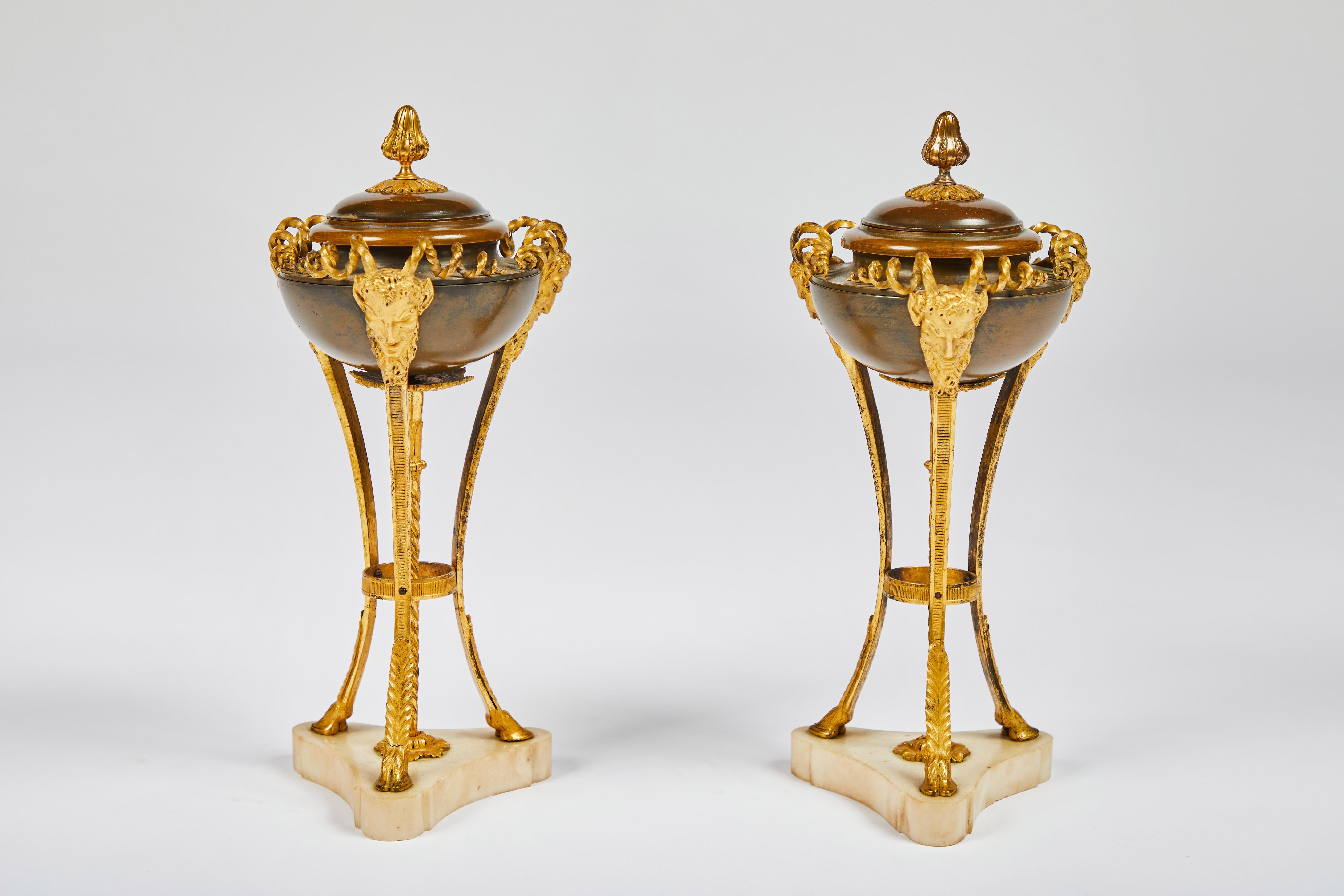 19th century, each comprising a shallow patinated bronze urn form and cover with gilt bronze finial, supported by three gilt bronze satyr masks with spiraling horns, terminating in hoof feet mounted on white marble base.
