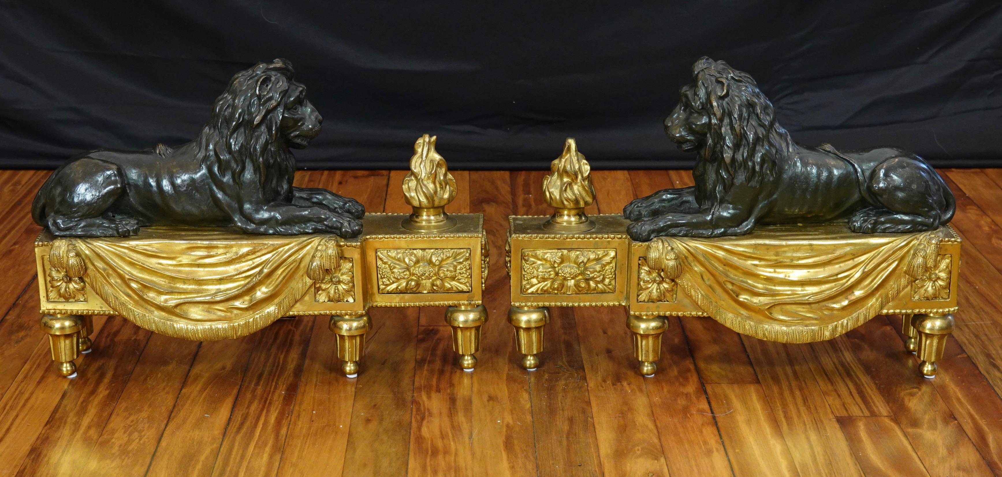 Pair of French Louis XVI style gilt and patinated bronze chenets or andirons featuring a reclining lion resting on a base with a flame, and ornamented with a fringed drapery, tassels, pearl beading, rosettes and other neoclassical details.  Each