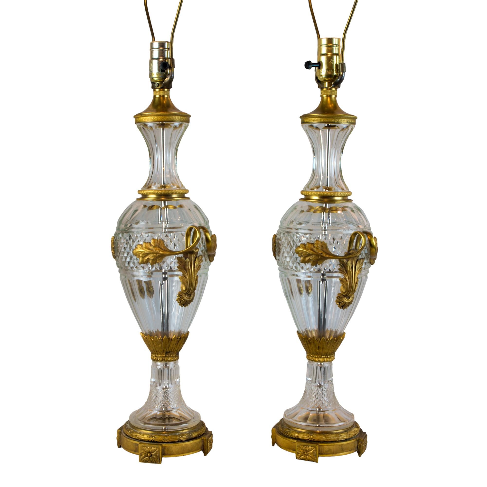 This lavish pair of Louis XVI Style Gilt-Bronze Mounted Cut Glass Two-Handled Table Lamps embodies the early 19th century spirit of grandiose decoration and inventive design.

Each lamp consists of an inverted baluster form vessel of cut crystal set