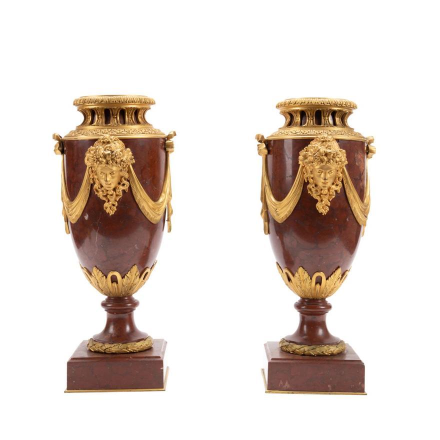 Paire d'urnes en marbre rouge griotte montées en bronze doré, de style Louis XVI, fin du 19e siècle. Elles sont ornées de masques féminins et de drapés, et reposent sur un socle. 
Dimensions : Hauteur 19