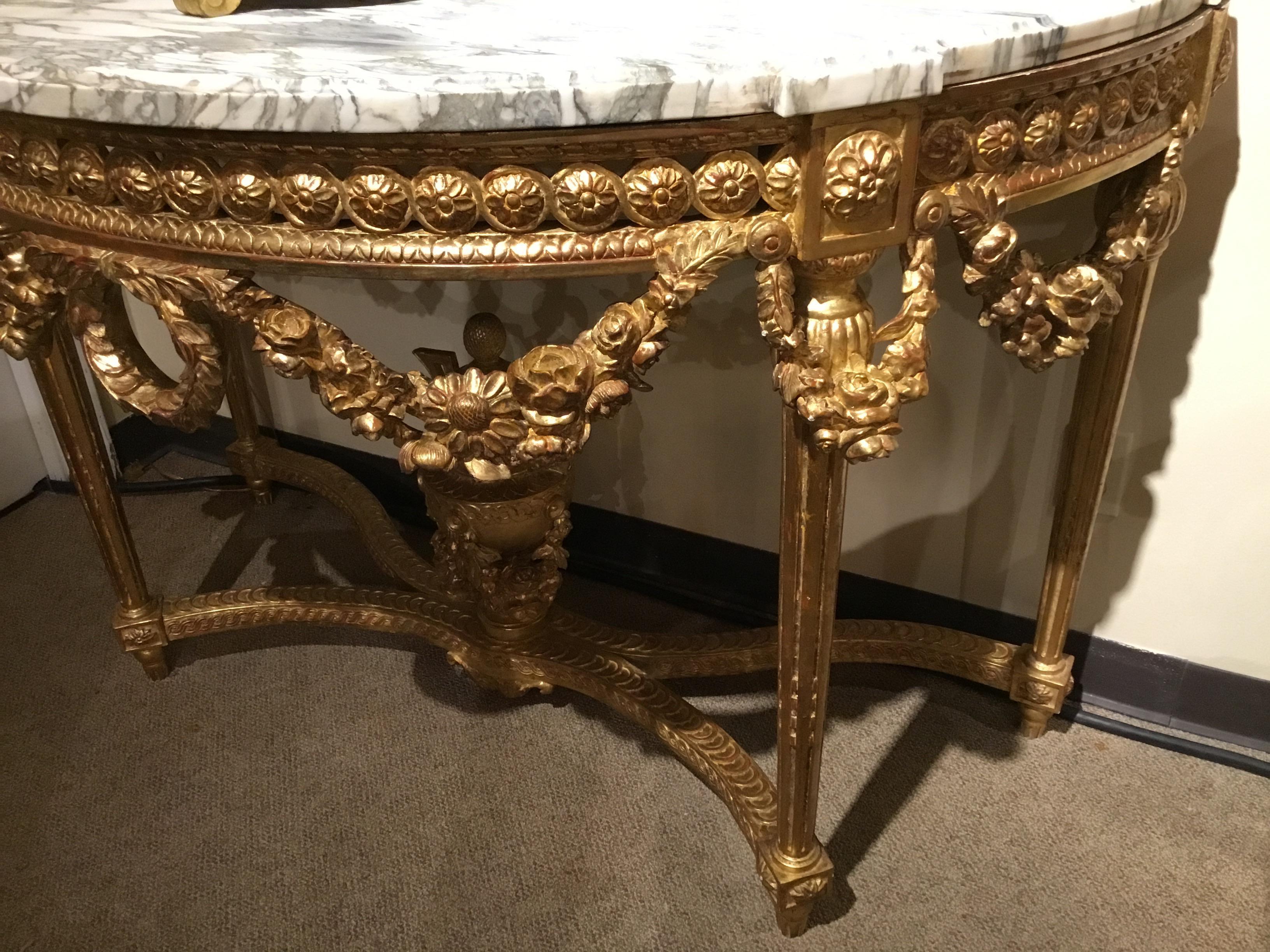 Demilune shaped consoles in giltwood having floral and foliate design in the draping and swags
that adorn the apron of these pieces. A stretcher in an X-formation comes together at the center
with an exquisite urn finial. The marble tops are in
