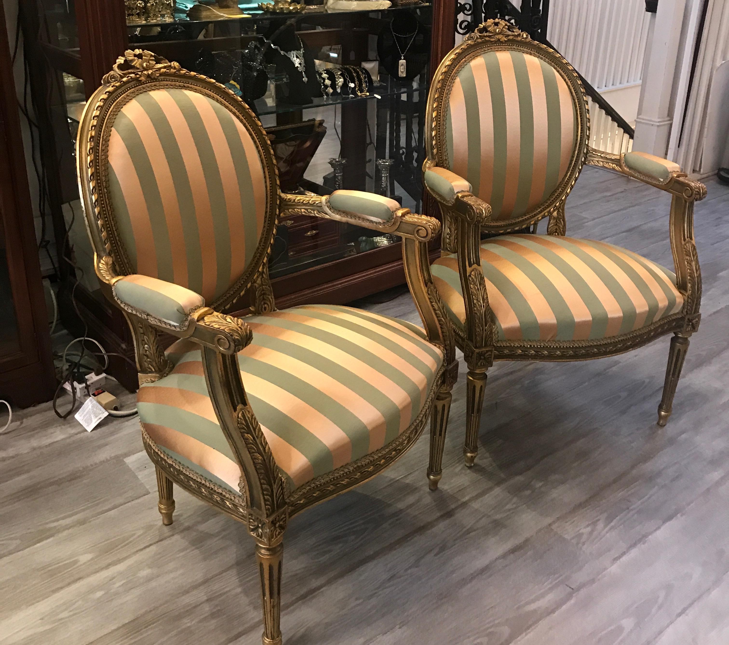 A pair of Louis XVI style giltwood armchairs. The oval backs with attached arms resting on four reeded and tapering legs. The new fabric in a gold and sage damask satin stripe, mid-20th century.