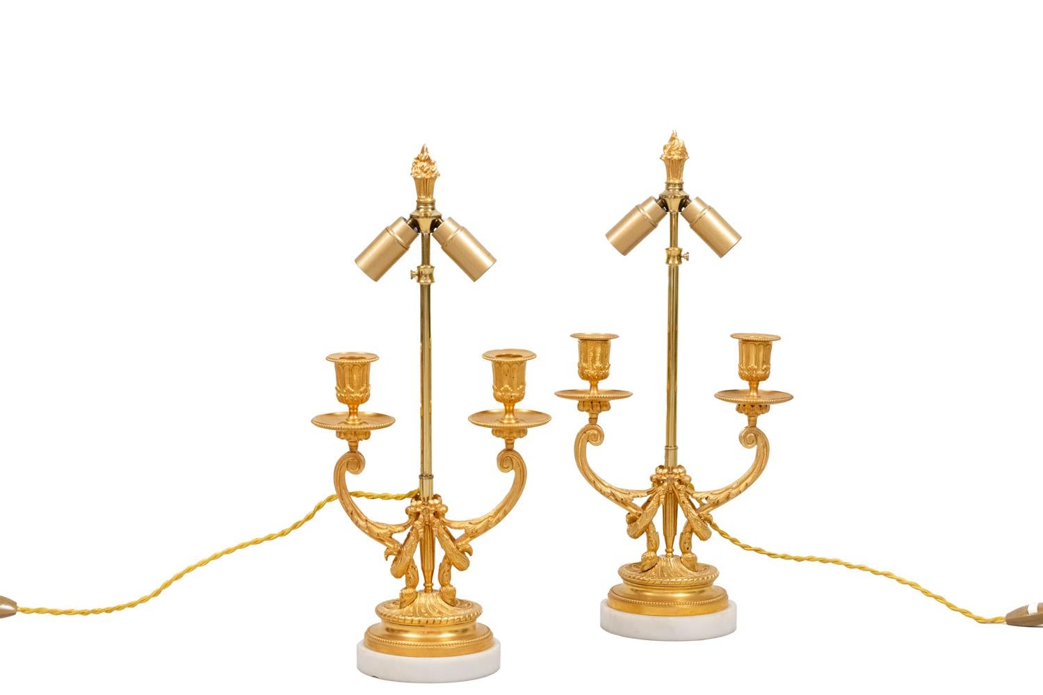 Pair of Louis XVI style lamps in gilt bronze. Two-arm lights in volute shape finished by hoofs and adorned with acanthus leaves. Bobeches decorated with flutes and leaves and cups adorned with a beads frieze. Shaft topped by a torch and decorated