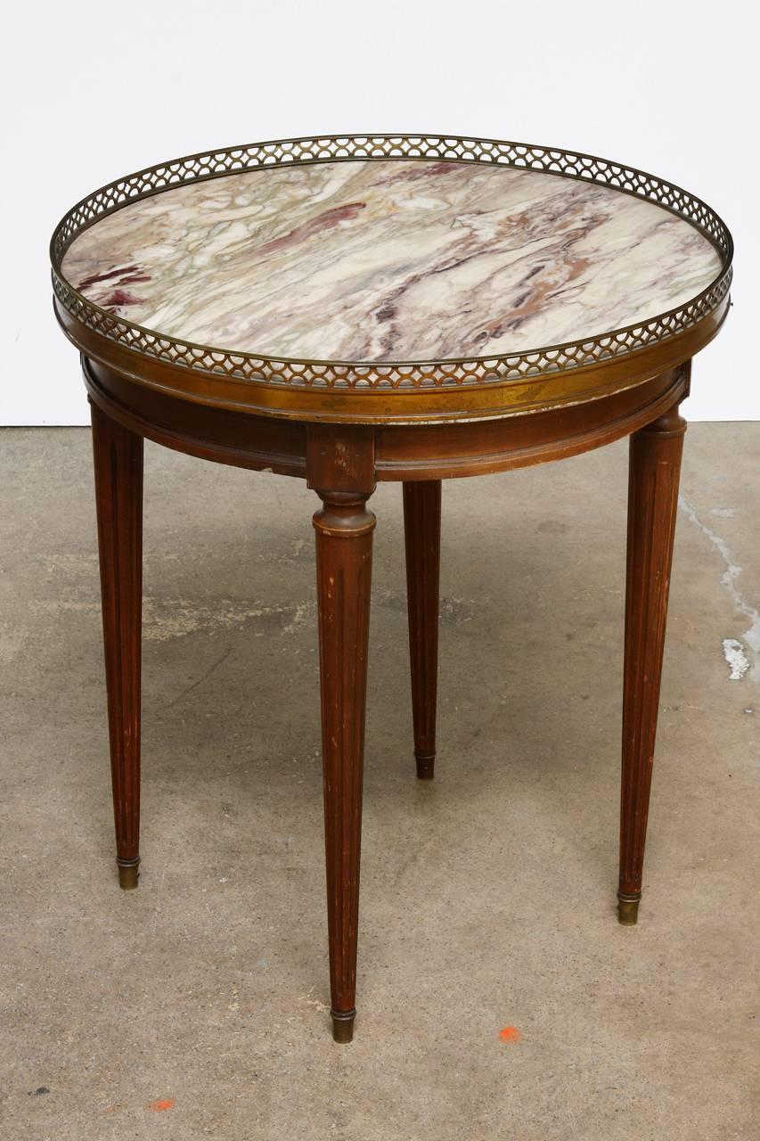 Classic pair of French Louis XVI style marble top gueridon drink tables. Carved from mahogany in the neoclassical taste. Each table encircled with a pierced brass galleried edge with one being 0.5” higher. The round marble tops are unique, one being