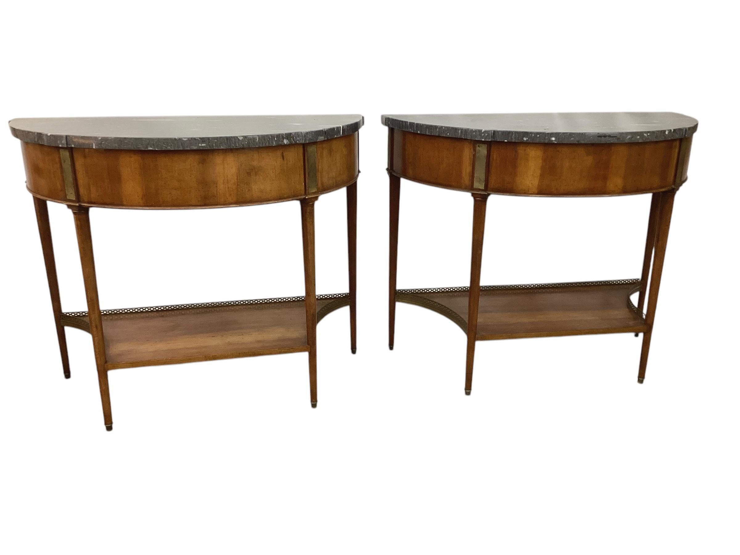Pair of Louis XVI Style Marble Top Mahogany Brass Mounted Demi-Lune Consoles. Each with a thick marble top and a lower shelf with a brass gallery. Each leg terminates in a brass sabot.
The tops were originally mahogany and had holes cut with a