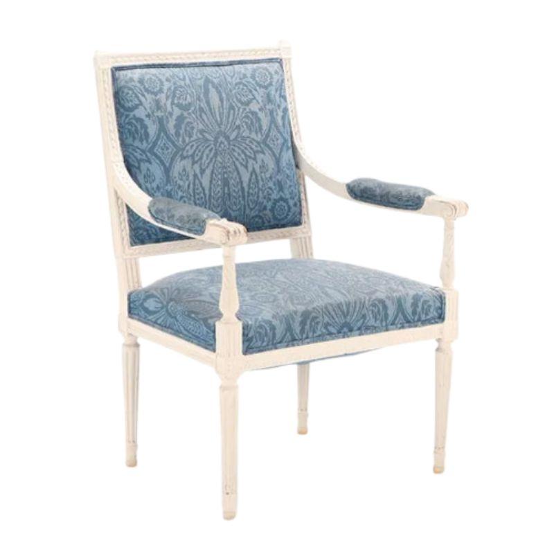 Pareja de sillas de comedor estilo Luis XVI pintadas y tapizadas, hacia 1930. Los sillones están tallados y pintados de blanco, con armazón de respaldo cuadrado y patas estriadas. Las elegantes sillas están tapizadas con tela de damasco azul de