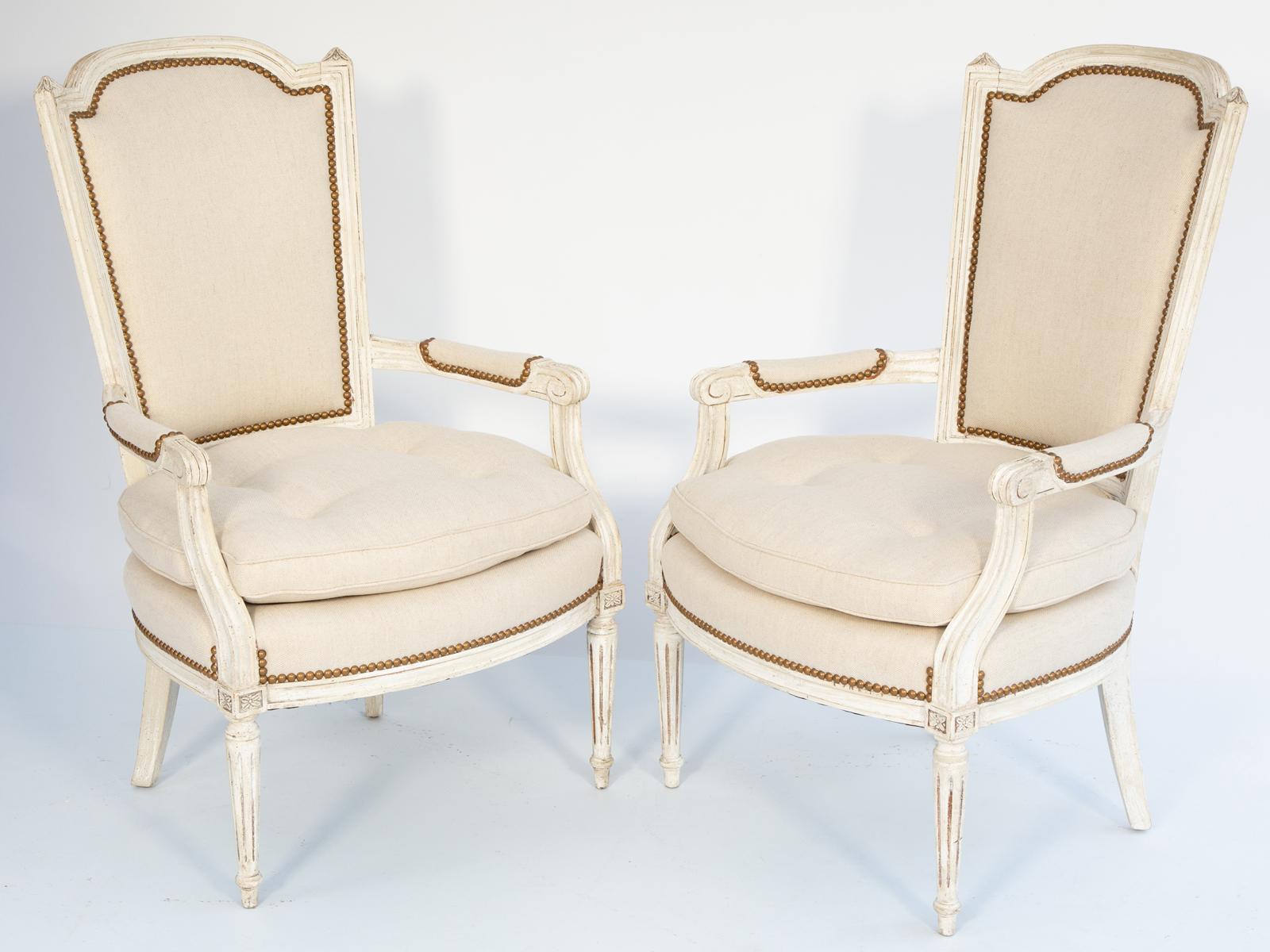 Paire de fauteuils avec une finition peinte montrant une usure naturelle, chacun ayant un cadre cannelé avec des arcs en forme de bouclier,  Le dossier rembourré, orné de fleurons en cassolette, se prolonge par des accoudoirs à enroulements et des
