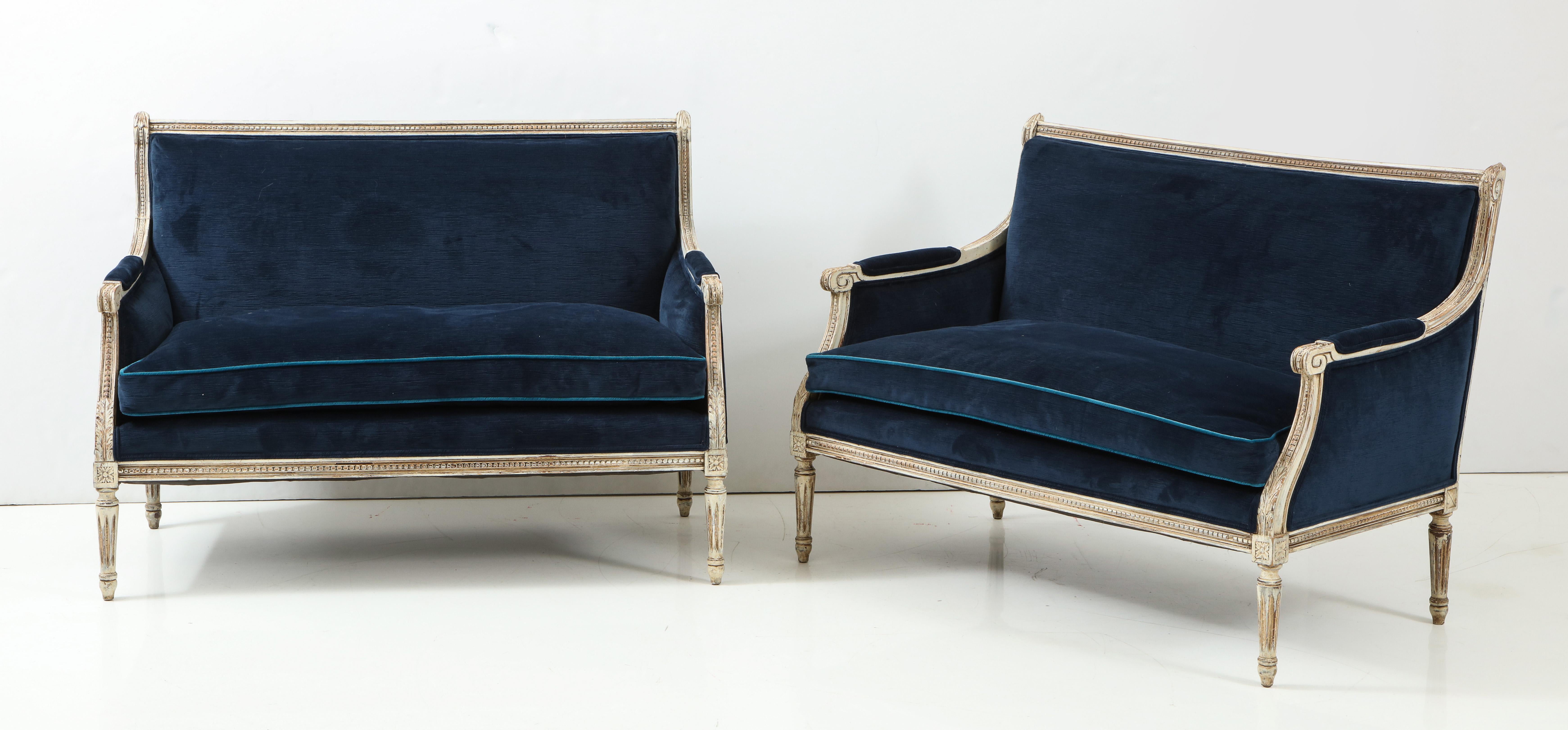 A pair of Louis XVI style painted settees covered in a rich navy velvet. The frames have beautiful carved detail, characteristic of the Louis XVI style. The finish is aged to a soft whitish-grey. The clean, Classic lines of these pieces make them