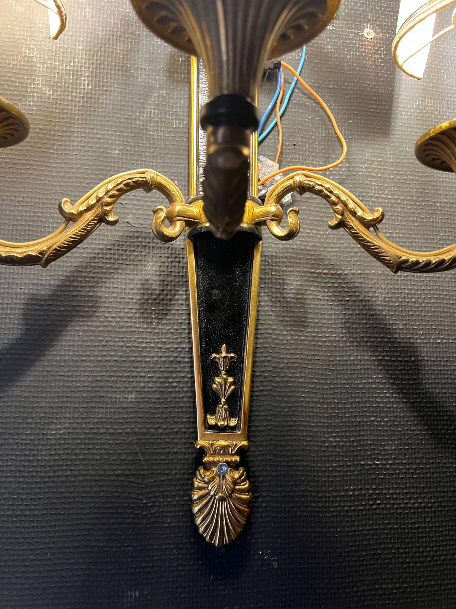 Elegante coppia di appliques francesi in stile Luigi XVI in bronzo patinato e dorato. Ogni applique presenta cinque bracci curvi decorati con foglie d'acanto e dettagli scanalati, sormontati da paralumi in tessuto plissettato. La piastra posteriore