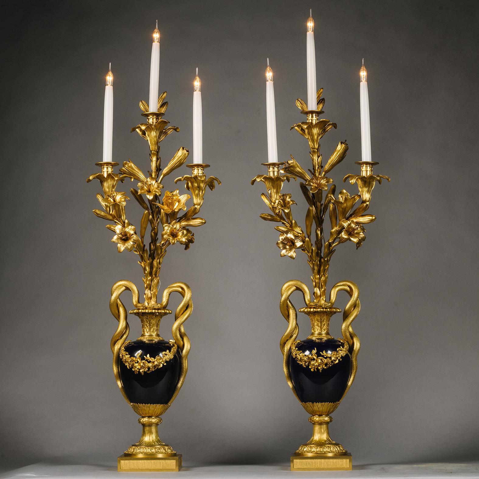 A Pair Of Louis XVI Style Gilt-Bronze And Royal Blue Sèvres-Style Porcelain Three-Light Vase Candelabra.

Each baluster-form vase applied with a fruiting swag, flanked by twin entwined-serpent handles, issuing lily flower branches with three candle