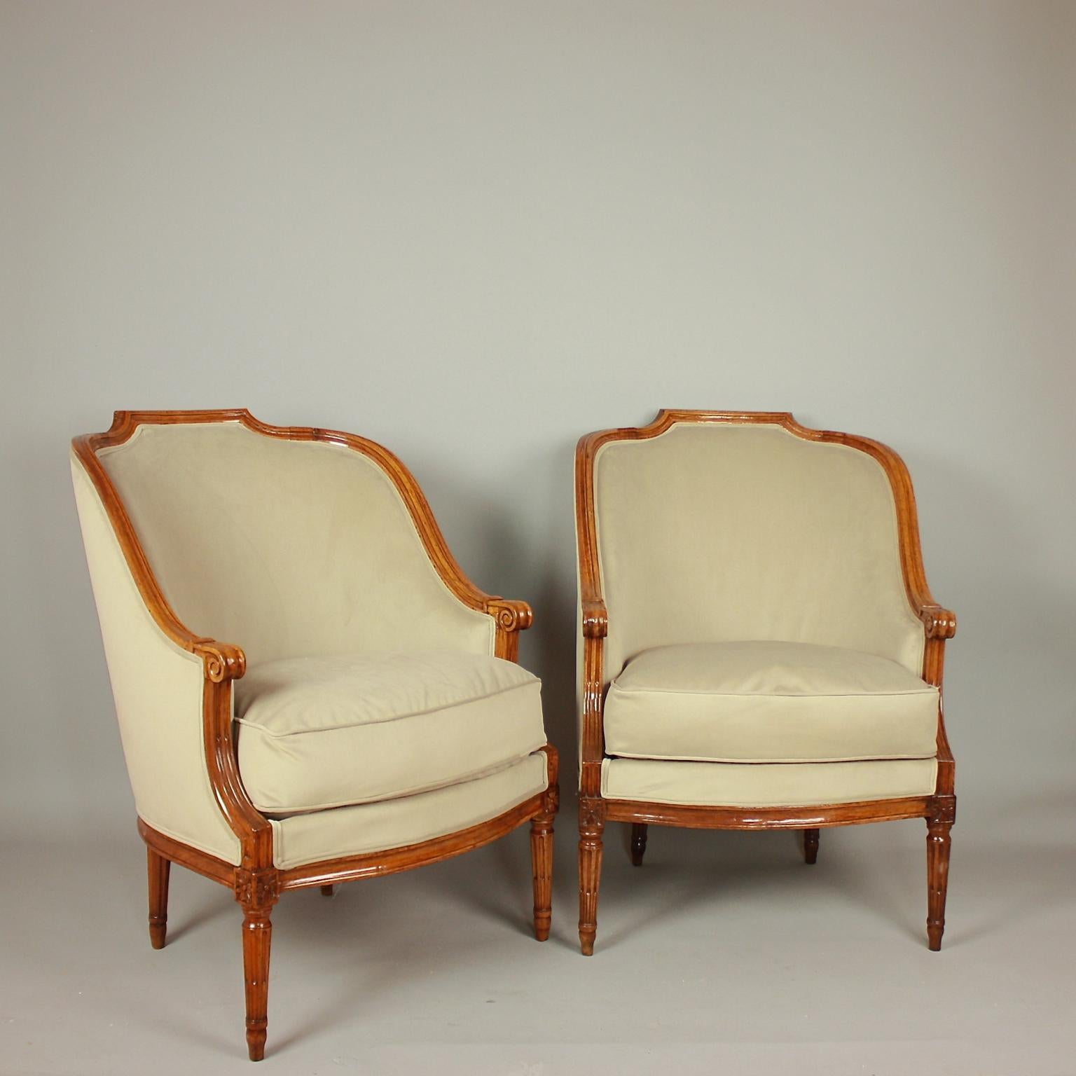 A pair of walnut bergeres or armchairs, each with a curved padded back. The top rail shaped to form a shallow arched and fluted top. The arm-rests descend to a spiral scroll to overlap the arm-supports, which sweep down again to meet the front legs.