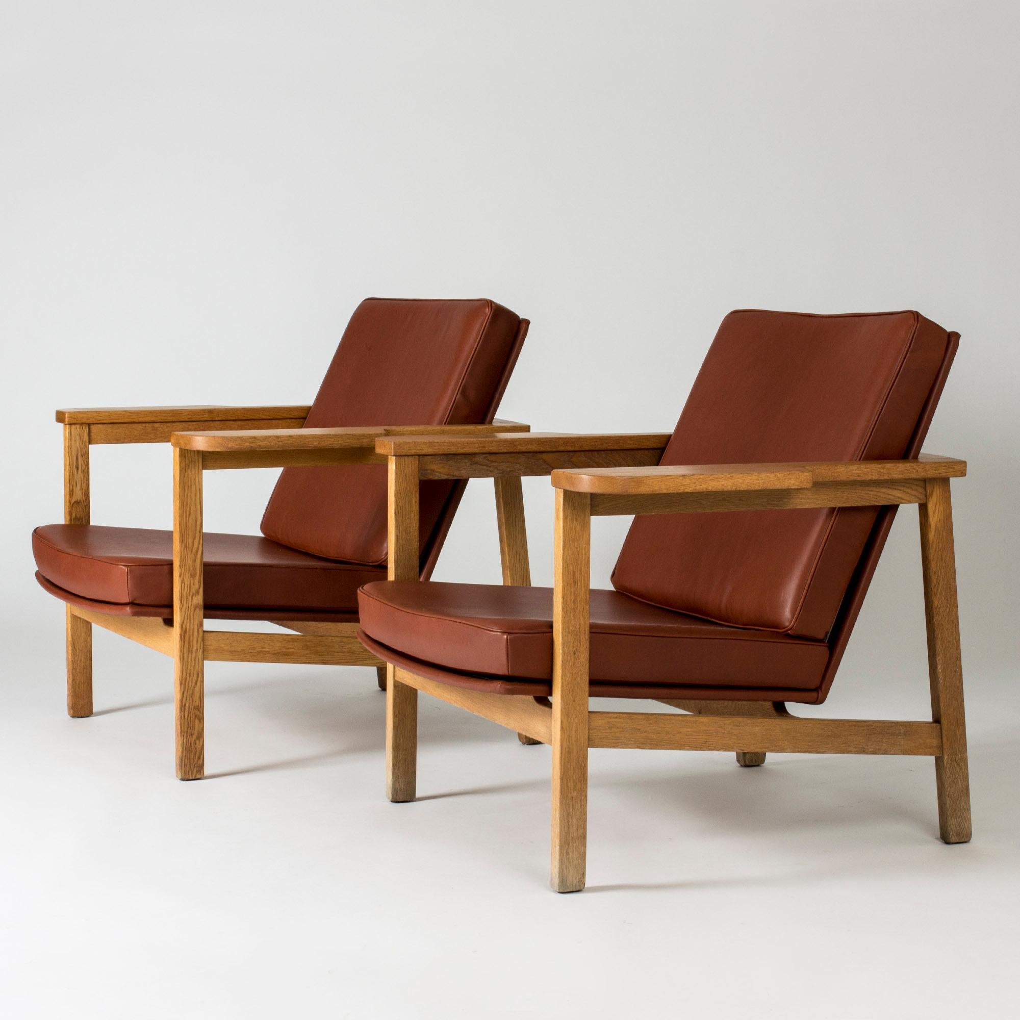 Pair of rare lounge chairs by Carl-Axel Acking in an imposing, luxurious design. Hefty oak frame with wide armrests and an elegant, open silhouette. Rust red leather upholstery.