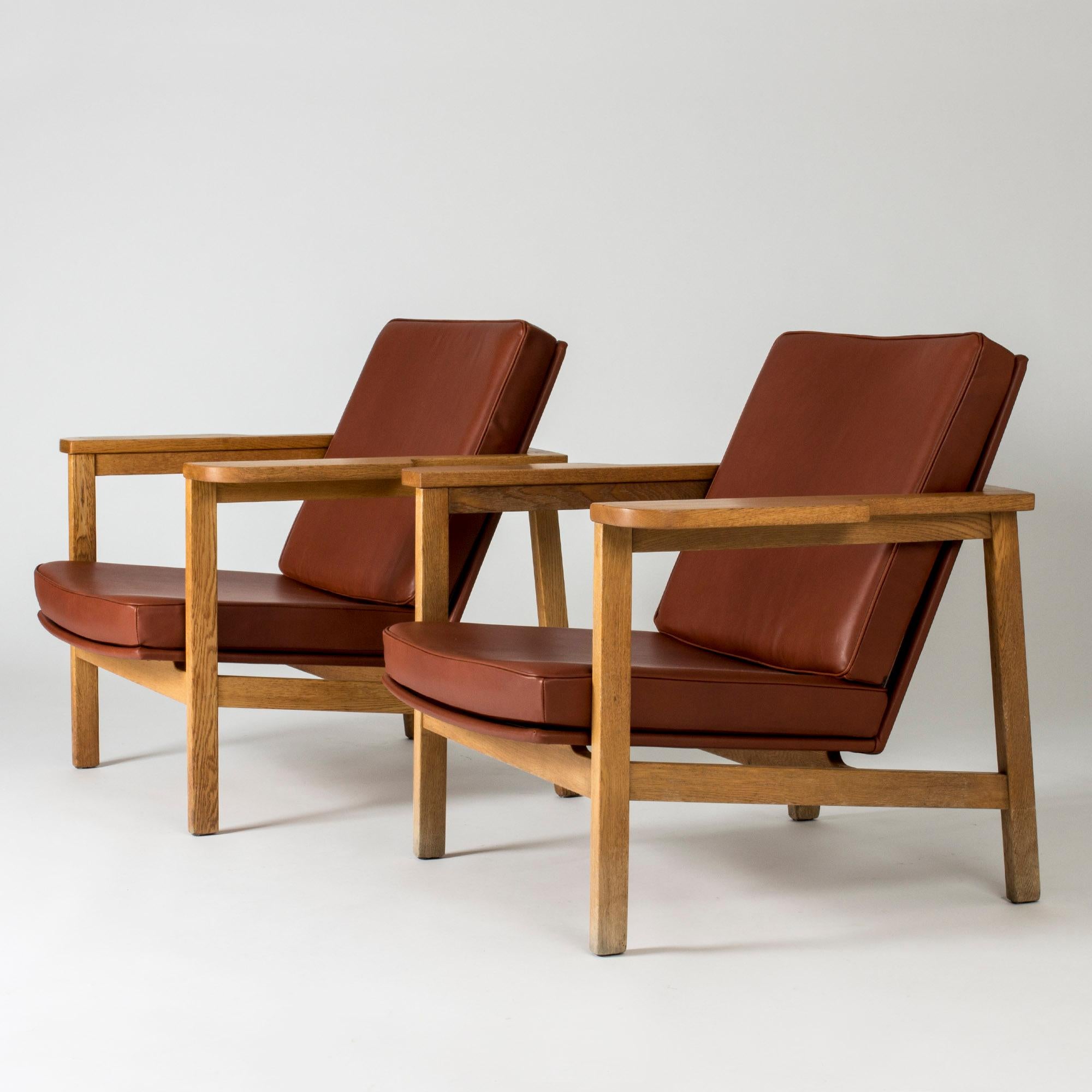 Paire de chaises longues rares de Carl-Axel Acking au design imposant et luxueux. Cadre en chêne massif avec de larges accoudoirs et une silhouette élégante et ouverte. Sellerie en cuir rouge rouille.