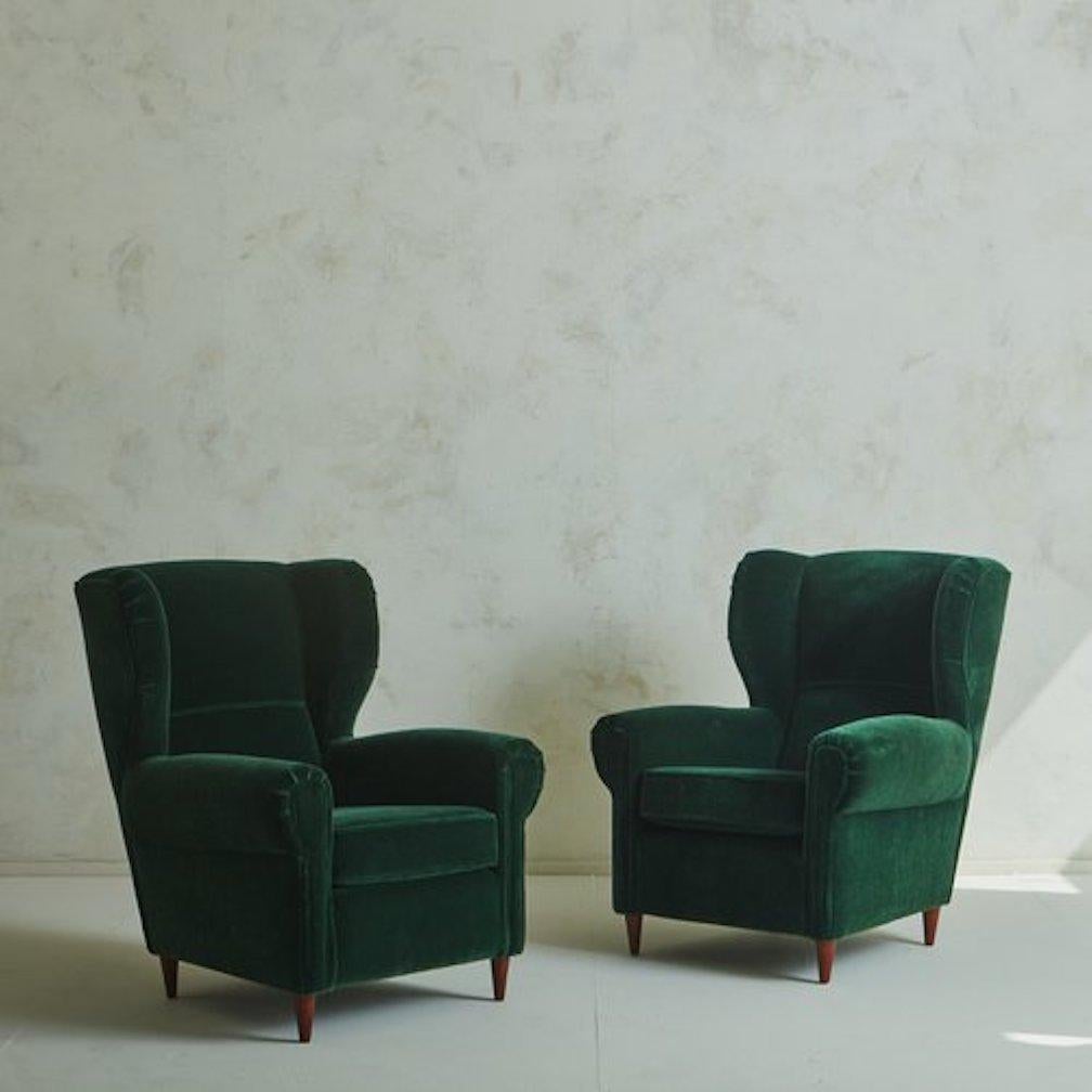 A pair of 1960s Italian wingback lounge chairs in the style of Paolo Buffa. These chairs feature tall, dramatic backs and stately arms. They were freshly reupholstered in an exquisite emerald green velvet with single welt detailing. The tapered