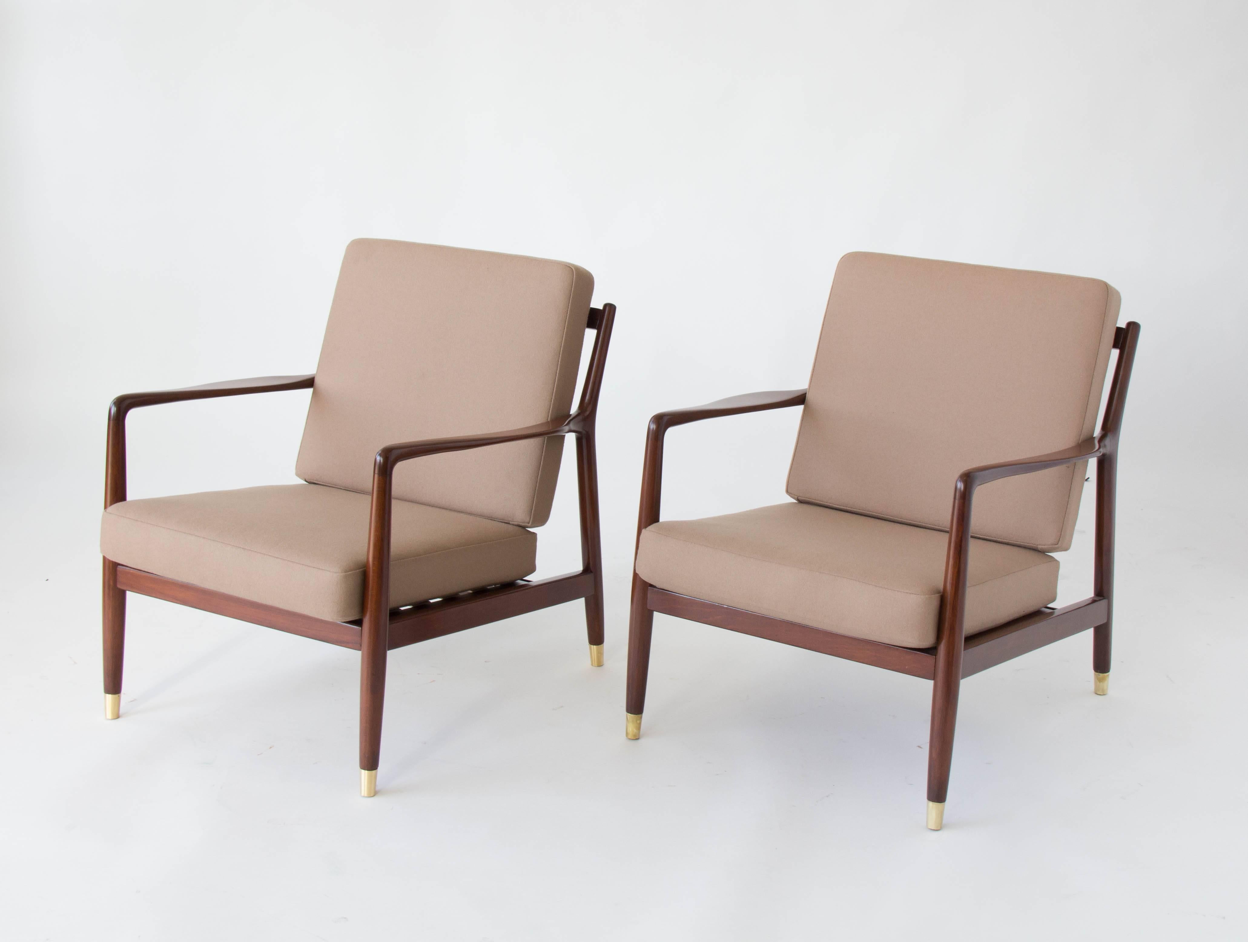 A pair of dark-stained teak lounge chairs designed by Folke Ohlsson and imported to the US under the DUX name. The chair has a deep seat framed by two delicately curved arms and a slatted backrest with a gentle angle. The cushions are covered in a