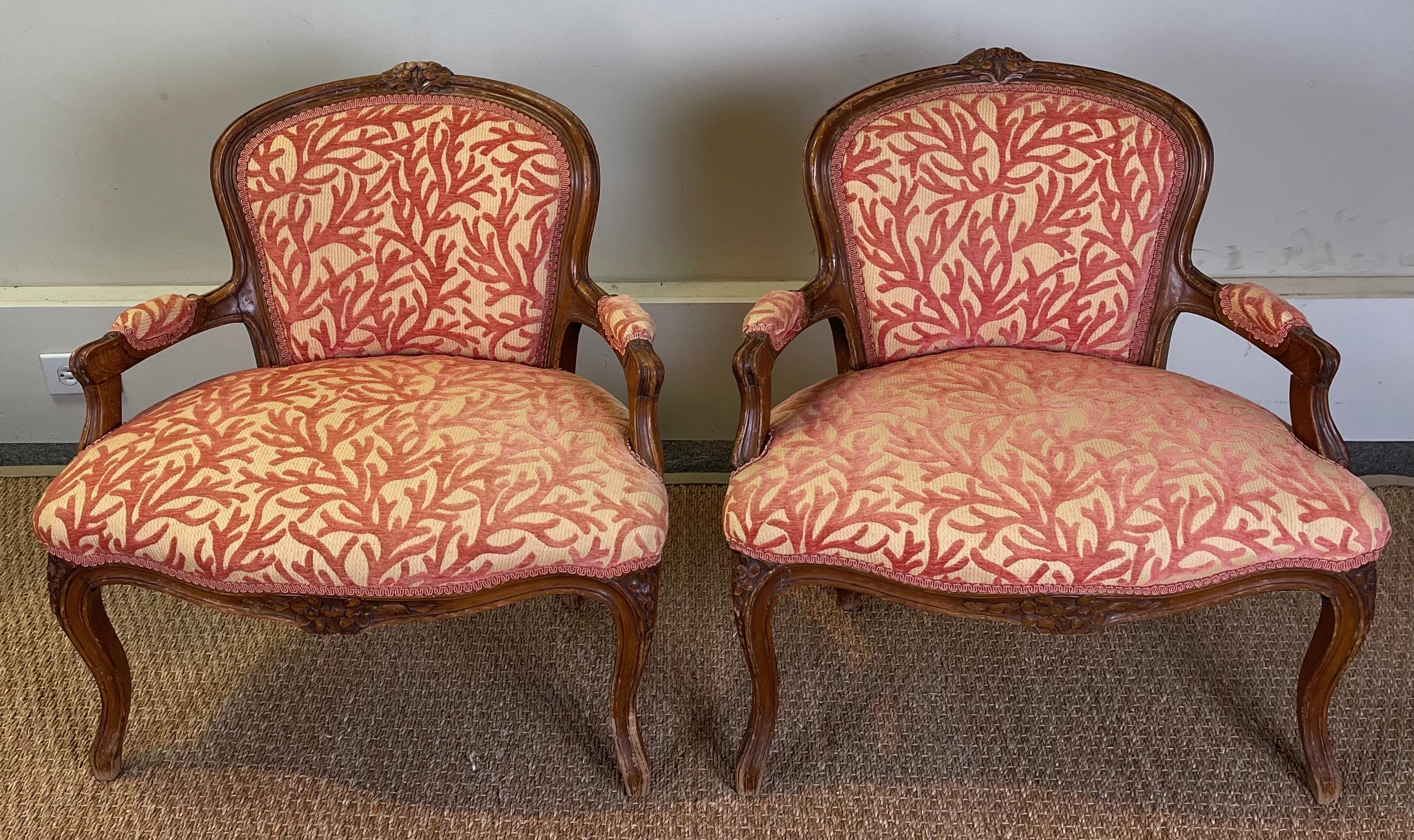 An unusual pair of early 20th C. carved walnut arm chairs or fauteuils with wide, low slung seats perhaps designed to capture the warmth of a fire covered in a coral branch velvet upholstery.