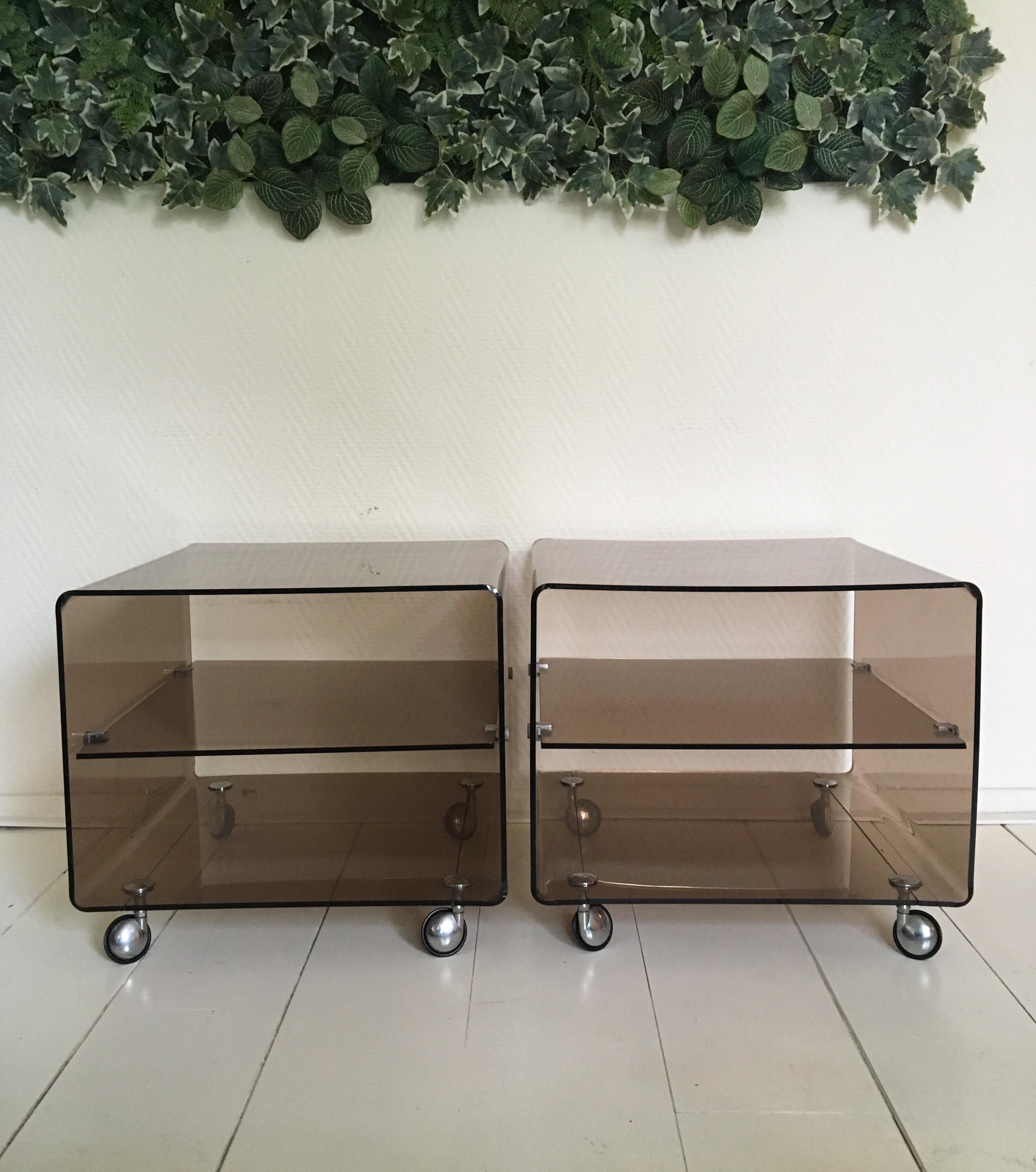 These Space Age Lucite side tables were designed by Michel Dumas for Roche Bobois, circa 1970s. Both tables feature a smoked brown/rose colored frame with wheels and a shelf. They remain in good condition but show some wear as scratches and