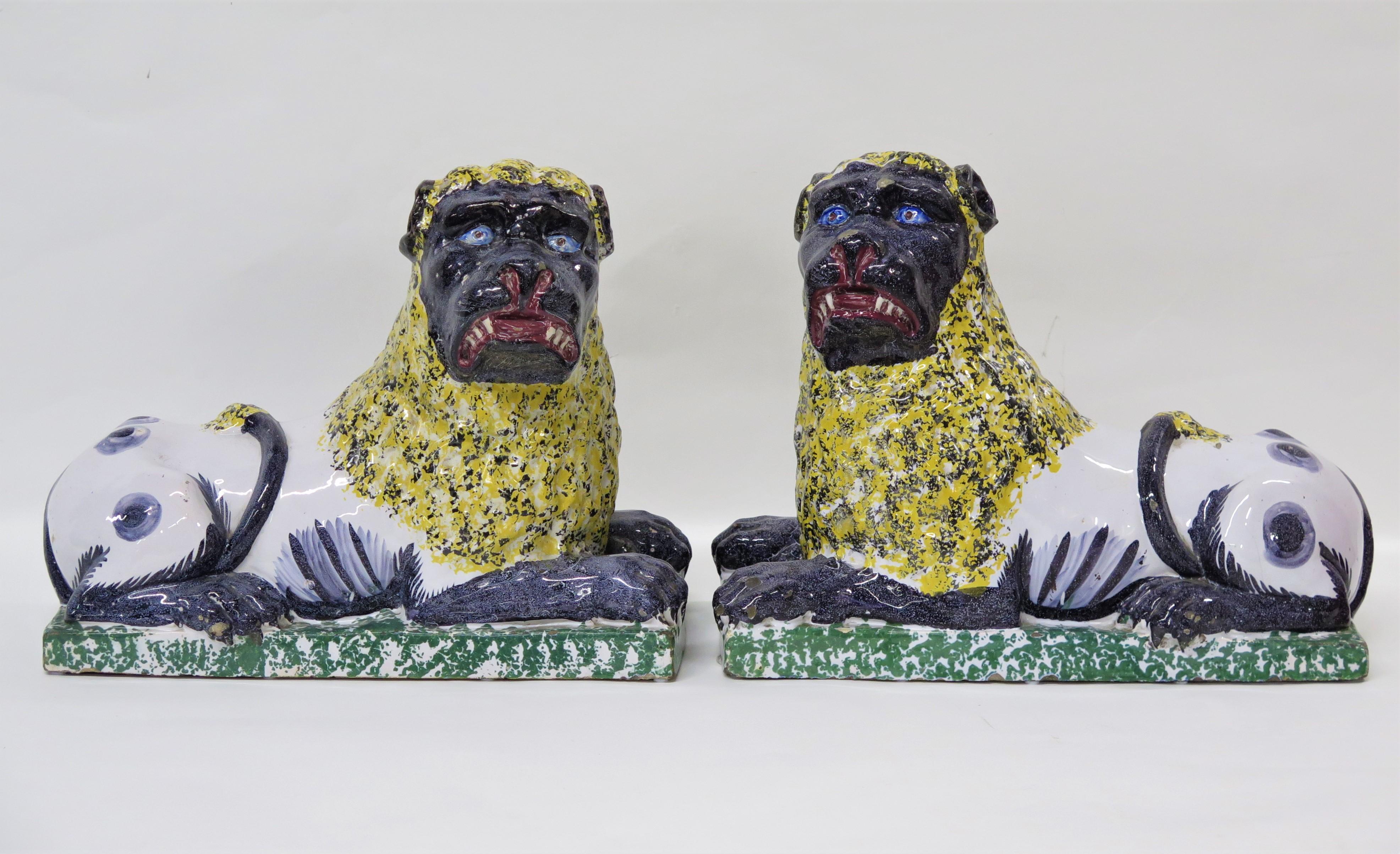 a large handsome pair of Lunéville faience lions, each shown resting on a green sponged plinth base with his tail curled over his back, their legs, paws, tails, and faces are painted gray-blue and their white bodies have gray-blue spots / markings,