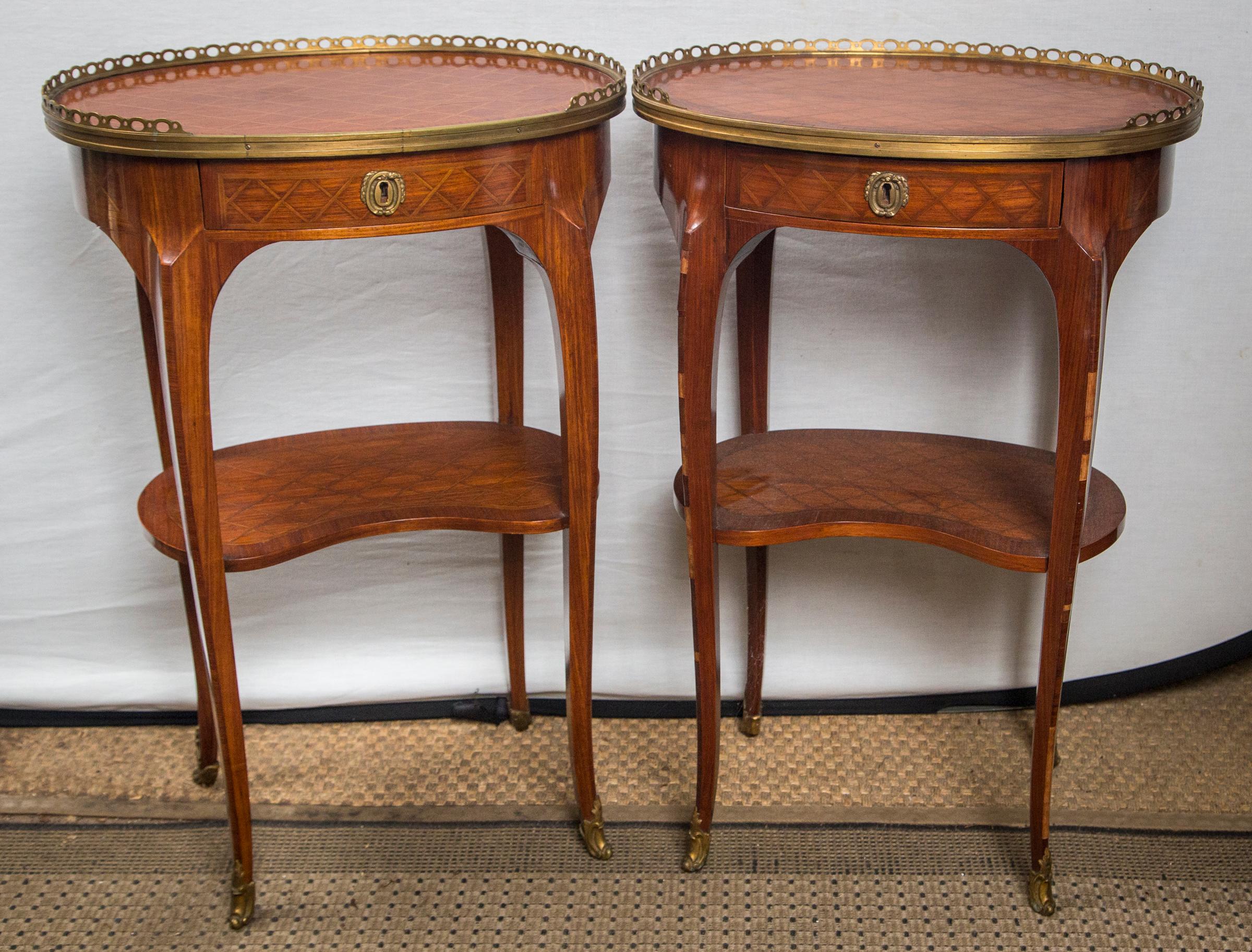 Of oval form with a brass gallery surrounding the cross hatch inlaid tops. One table having slightly lighter inlay of the cross hatching. The lower shelf of kidney shape. Each has a single drawer. Graceful transitional style legs, ending in brass