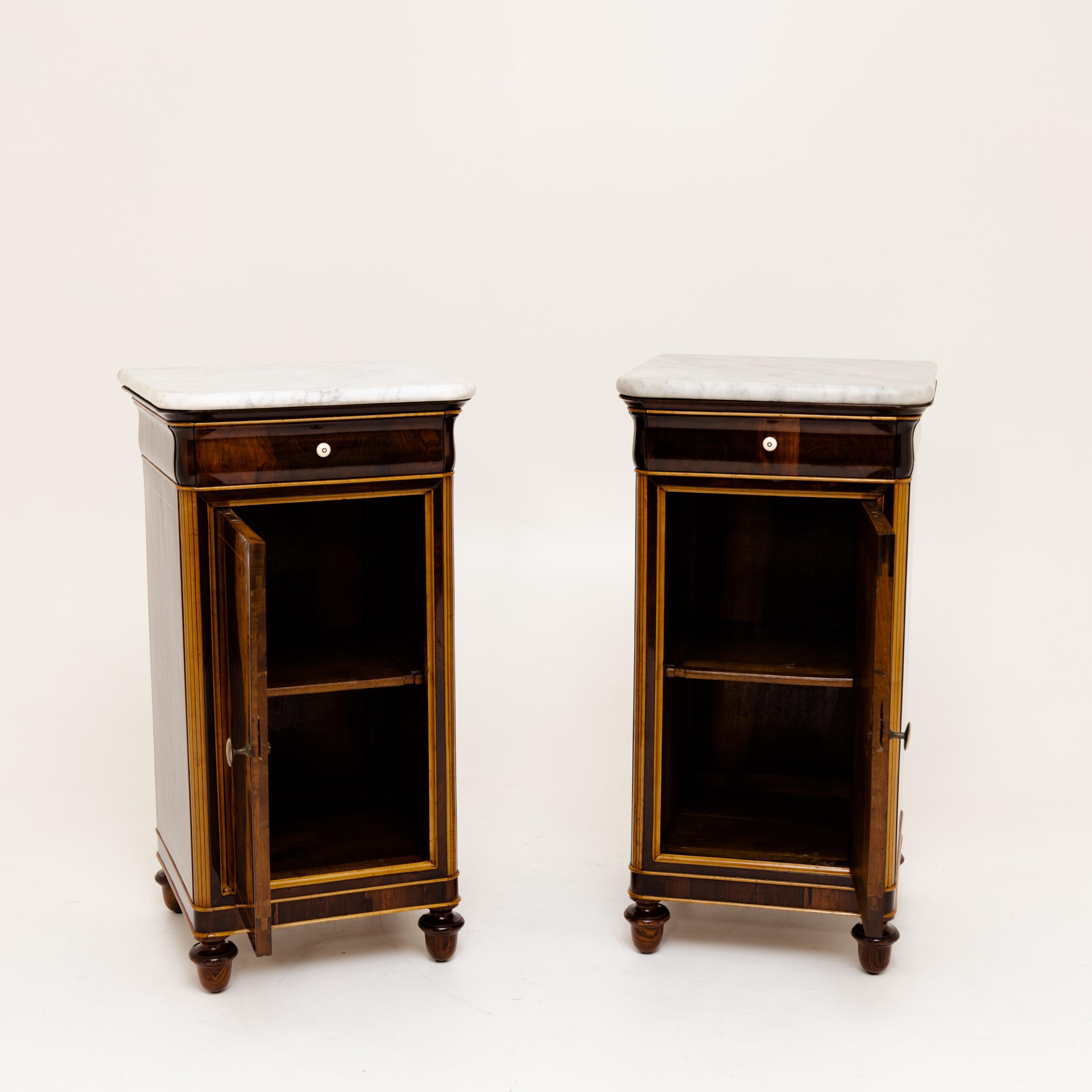Pair of oppositely designed bedside cabinets on conical legs with one door, one drawer and a marble top. Mahogany and maple veneered.
