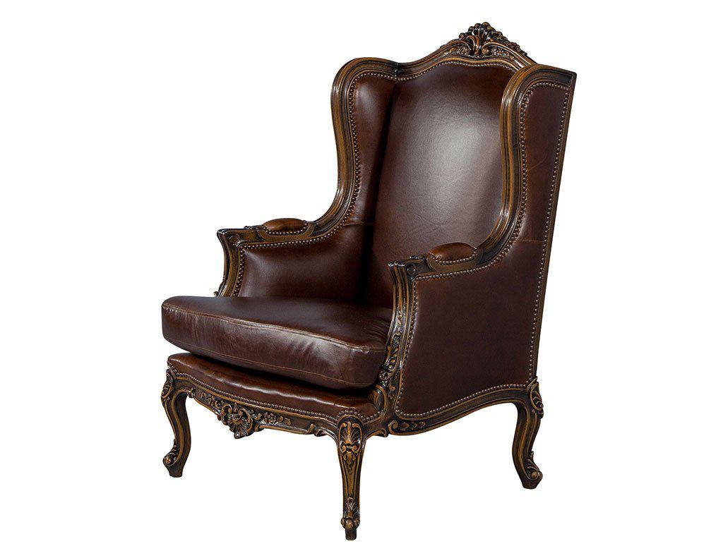 A set of two stunning wing chairs fashioned in the style of the Louis XV bergère, with a carved mahogany frame, high back and flared seat with a gently serpentine front. Full brown leather upholstery trimmed with head to head nails complements the