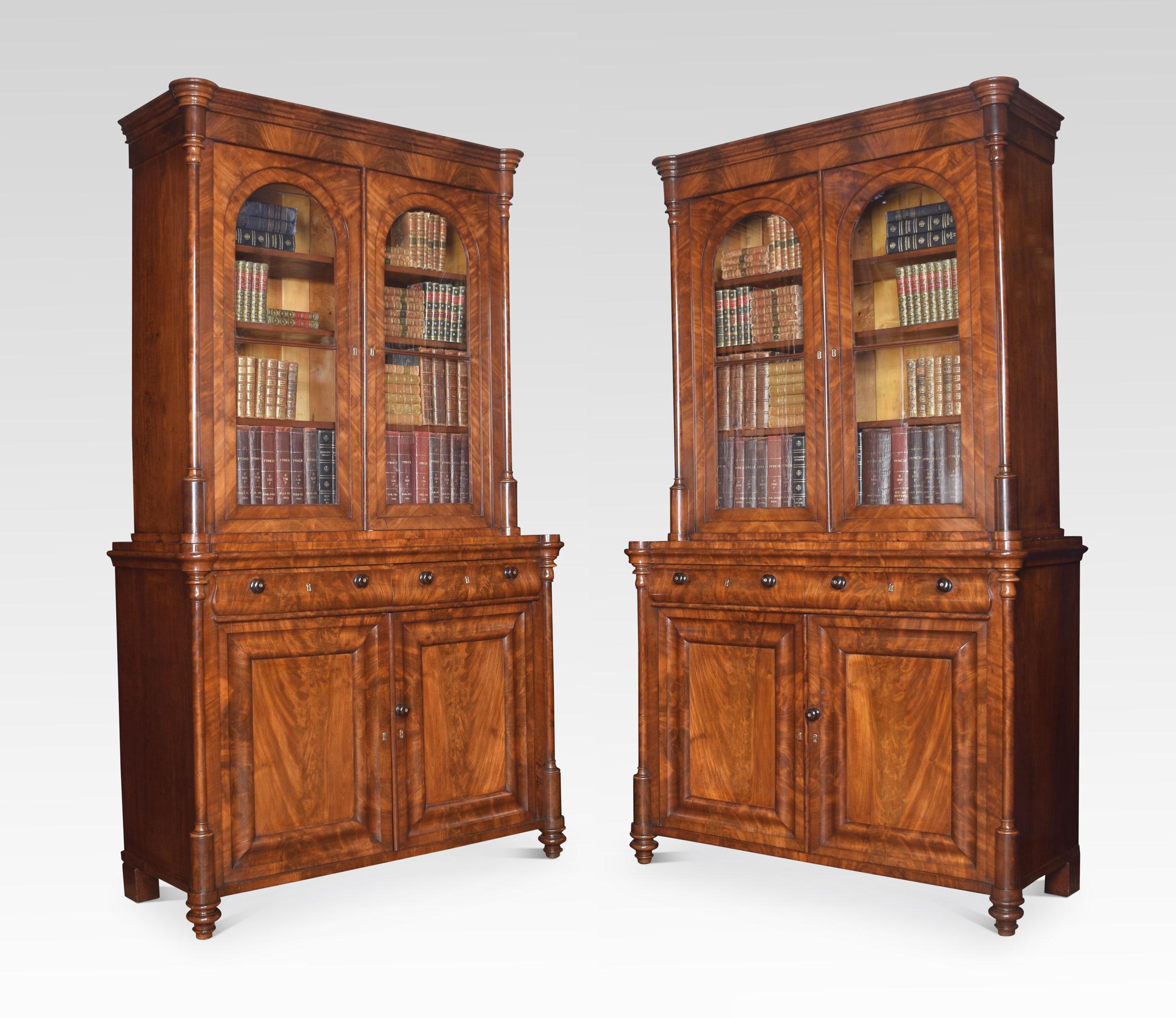 Pair of mahogany library bookcases, each with moulded cornice, above a pair of glazed panelled doors enclosing adjustable shelved interior. Flanked by three-quarter round turned pilasters.
To the base section fitted with two freeze drawers with