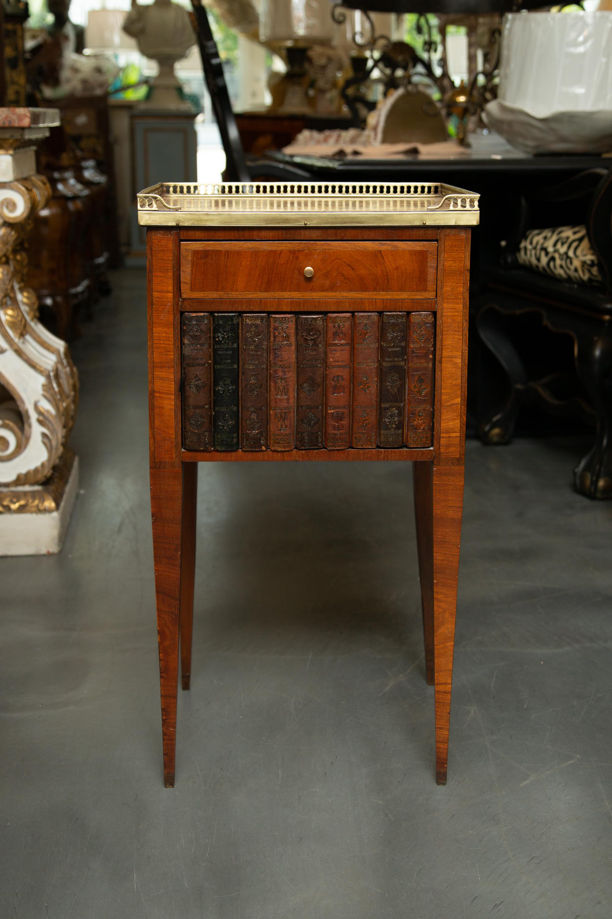 This is a charming pair of inlaid Louis XVI style side tables. The tops are aggregate stone. They are framed by a brass pierced gallery over a central drawer and cabinet door faced with book spines, early 20th century.