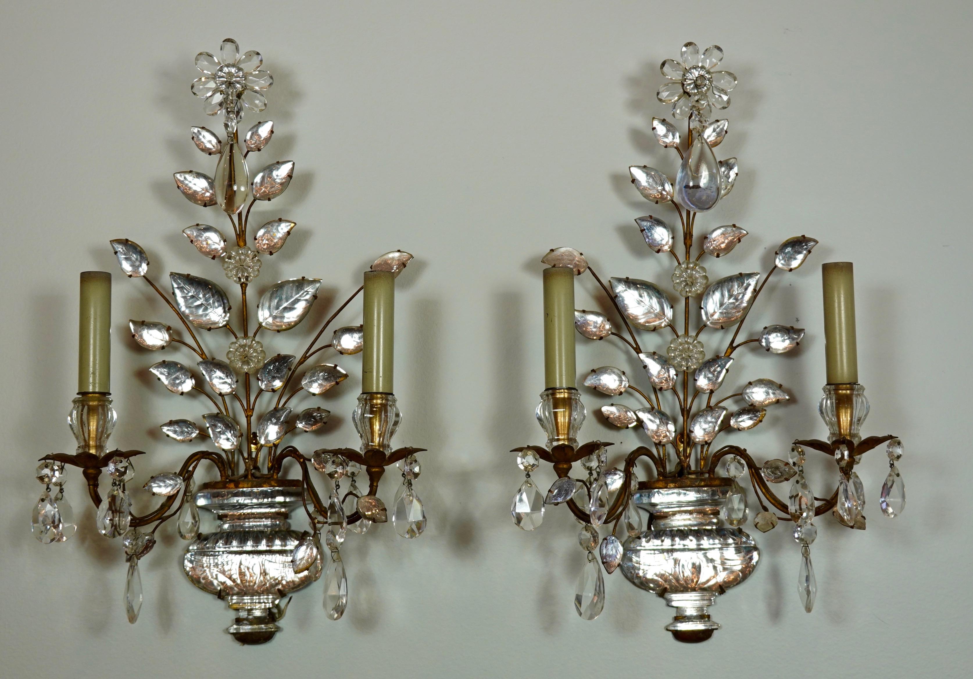 A pair of French gilt-iron and crystal two-arm sconces by Maison Bagues (mid-century). The sconces feature a hand-strung flower and spray of leaves planted in a crystal urn base. Crystal drops are suspended from the flower, bobesches and arms.
