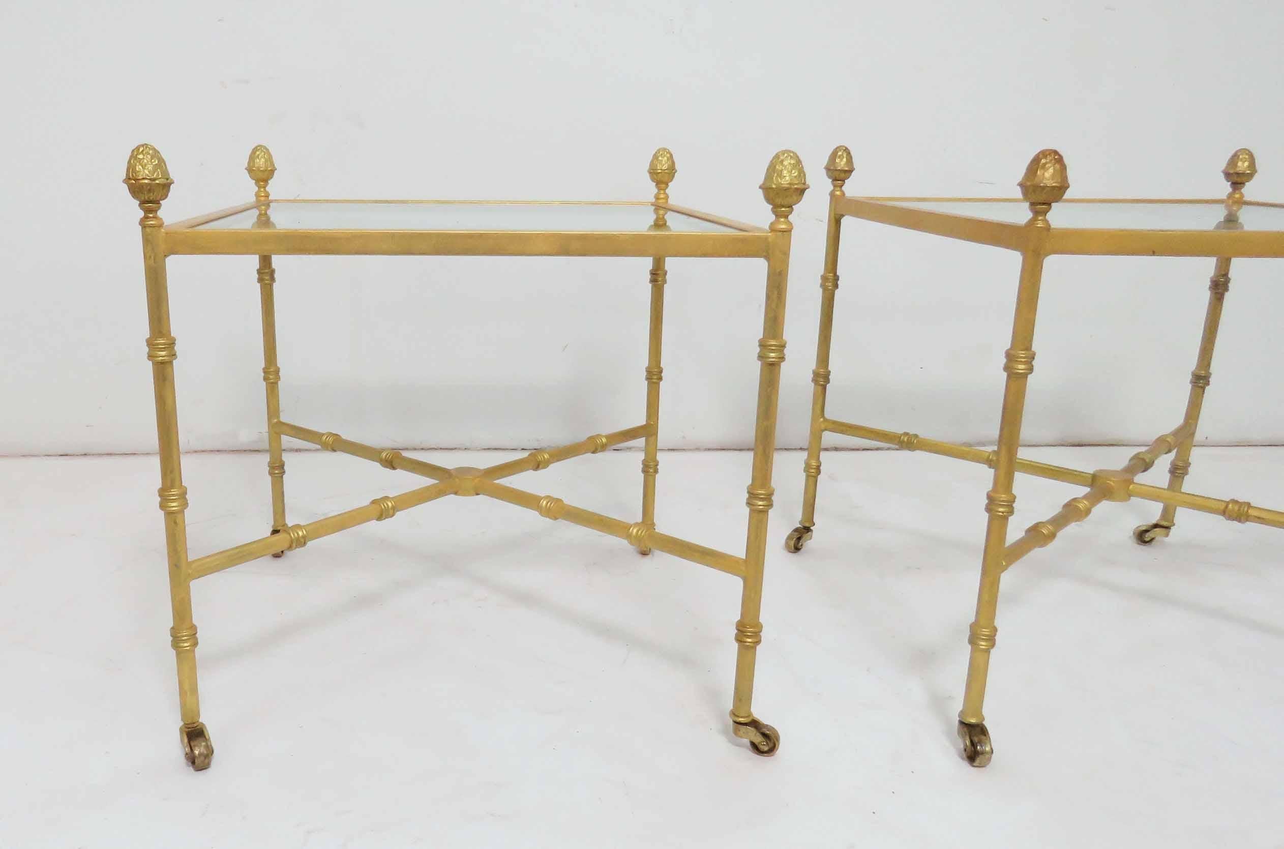 Pair of glass top end tables in gilt metal with acorn finial accents, X-form stretchers, and delicate solid brass casters.

Measures: 20.75