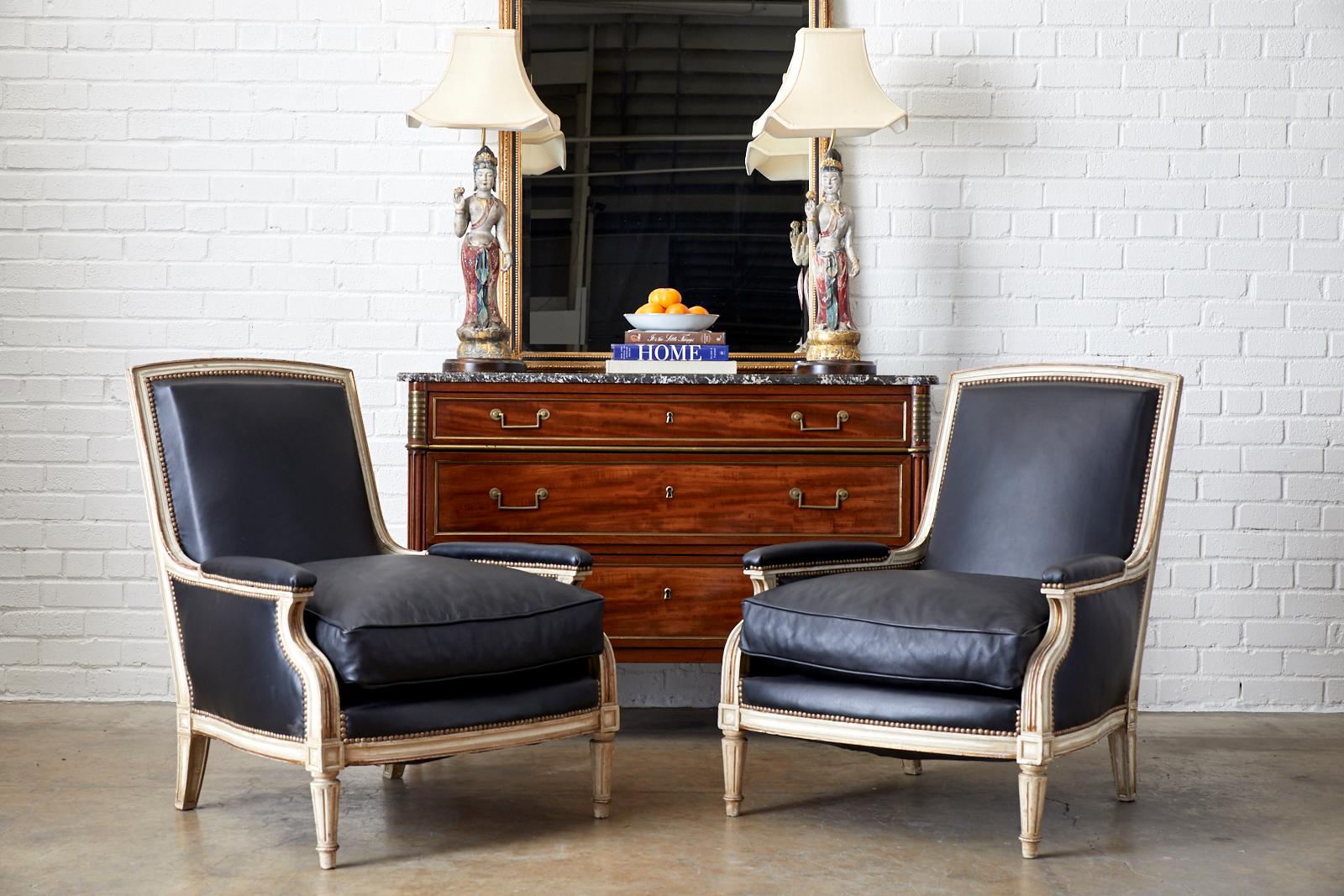 Fantastic pair of French painted bergere armchairs made by Maison Jansen. The large chairs feature a parcel gilt aged lacquer frame with a square flat back. Lavishly upholstered with thick, supple black leather hides having brass tack nailhead trim