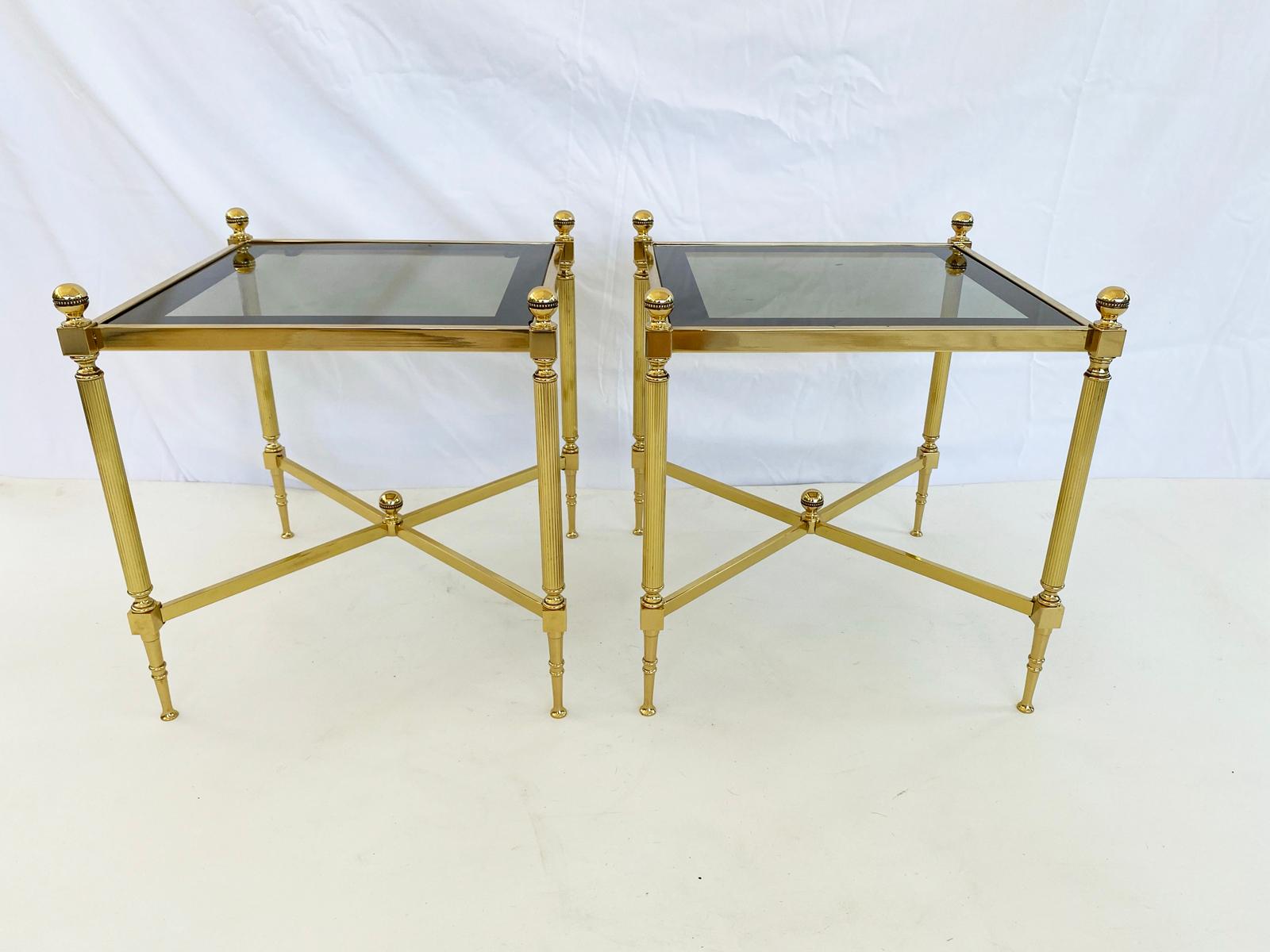 Pair of end tables, of polished brass, each holds their original smoked glass tops with mirrored border, a round beaded-ball finial adorns the top of each reeded leg, joined by an X-stretcher, centered by matching finial, raised on tapering cone