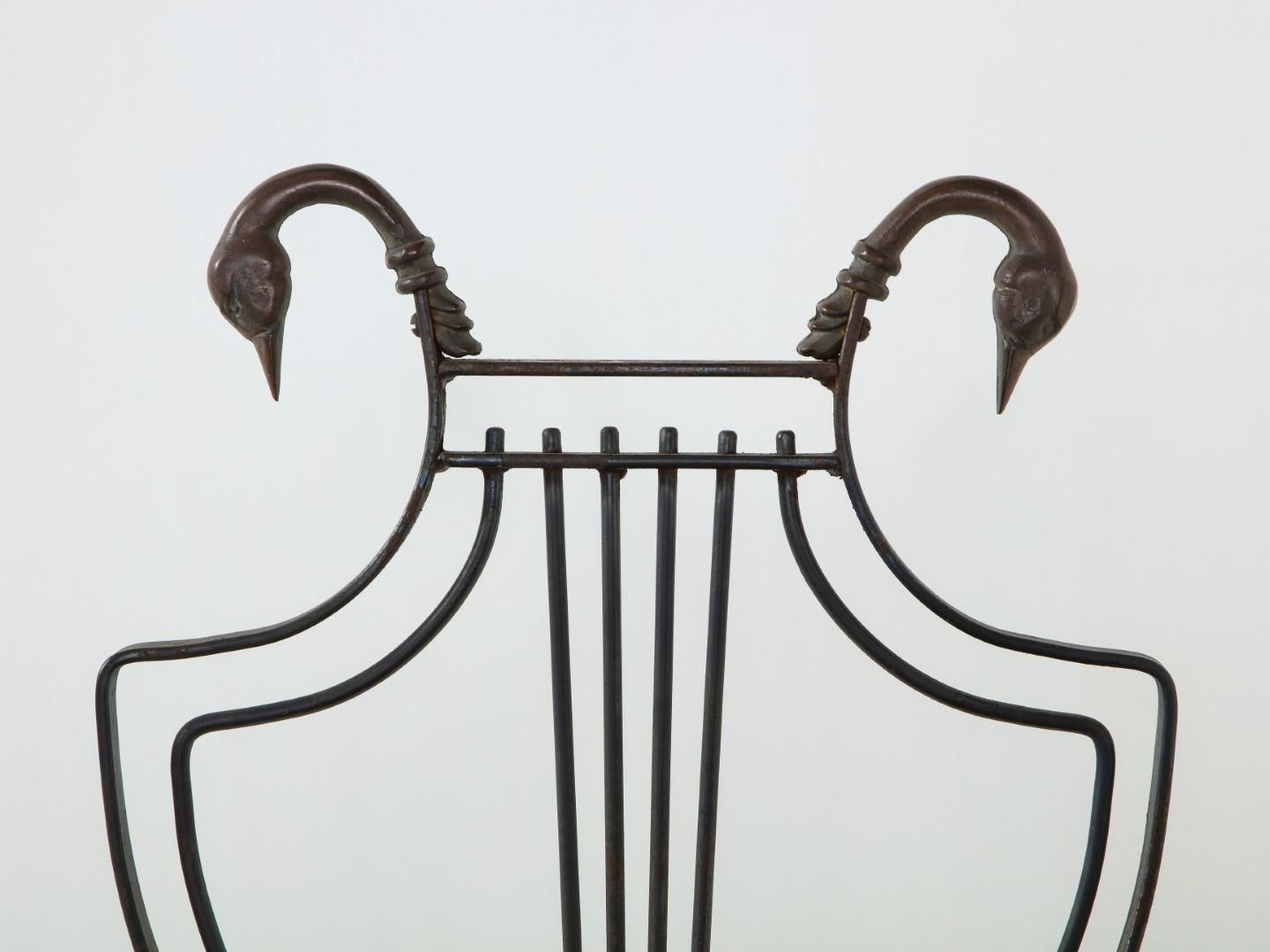 A pair of Maison Jansen vintage black wrought iron and brass lyre-back arm chairs, with paw feet. Swan head accents at tops of backrest left and right. Patina from outdoor use, cost considers the need to reupholster.