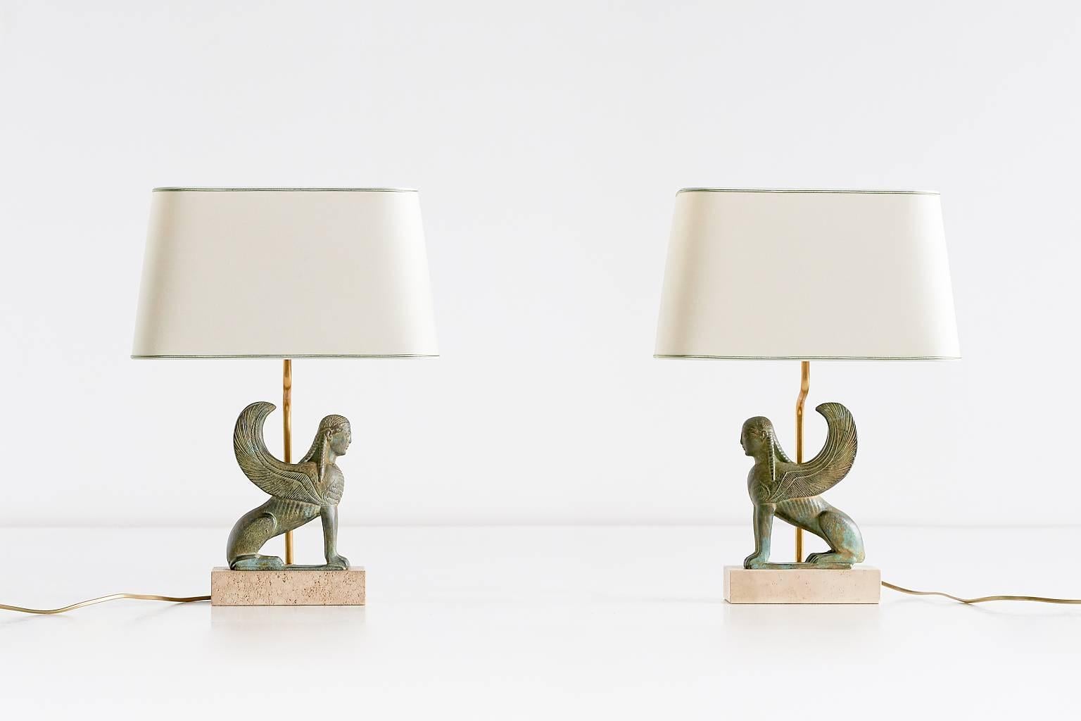 This striking pair of table lamps was designed and produced by Maison Le Dauphin in the 1970s. The intricately decorated golden green ceramic sphinx figures stand on a travertine base. The new ivory shades with a subtly contrasting green trim detail