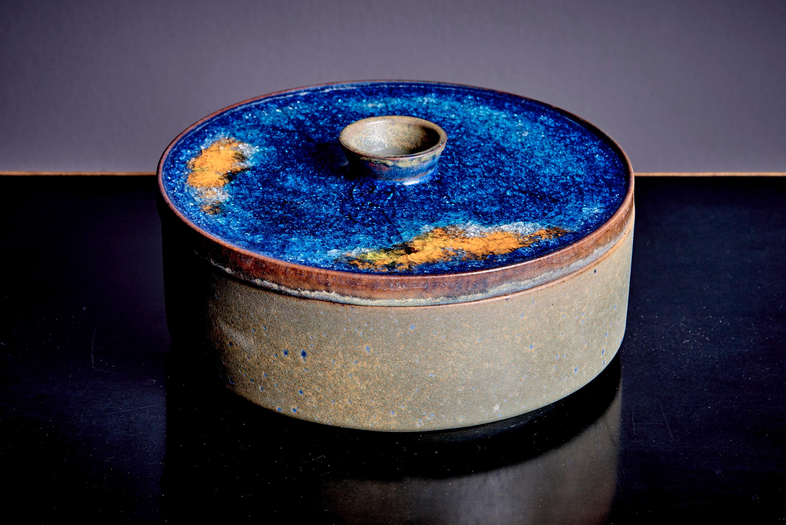 Nice pair of 1960s Majolika ceramics, known for bold glazes and artistic designs. The larger lidded container has a textured cobalt blue lid with gold and orange accents, contrasted by its earthy-toned base. The smaller spoon-like dish has a glossy