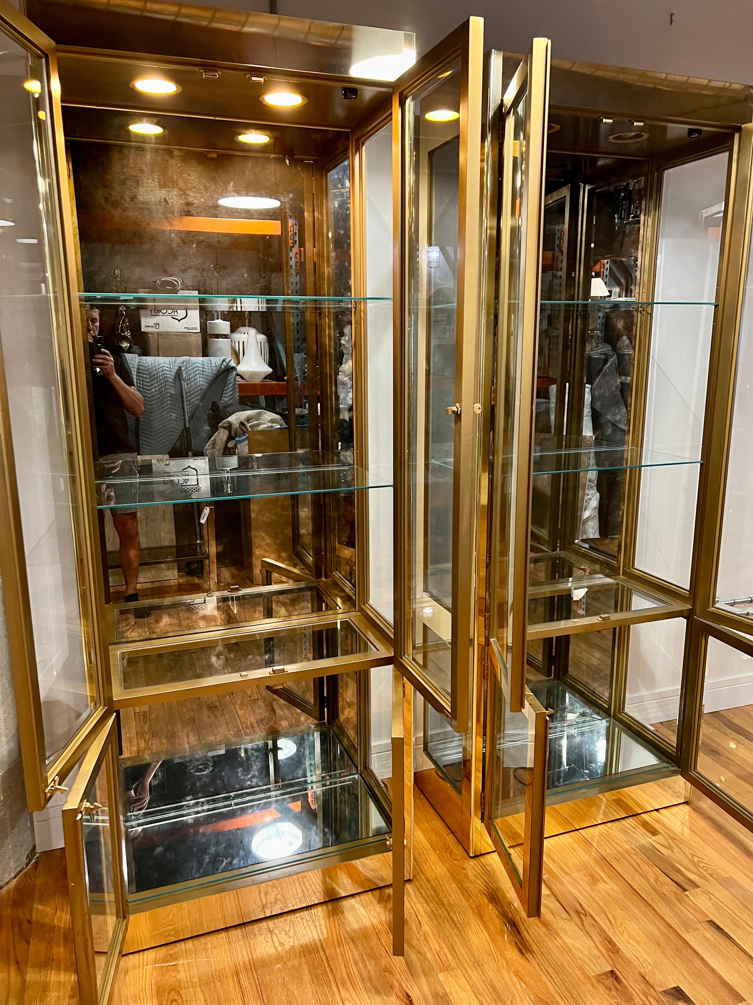 Pair of brass and glass display cabinets or vitrines by Mastercraft. These chic and elegant cabinets are in remarkably good condition for their age featuring a smoke mirrored back panel and four glass shelves. The cabinets can be illuminated and